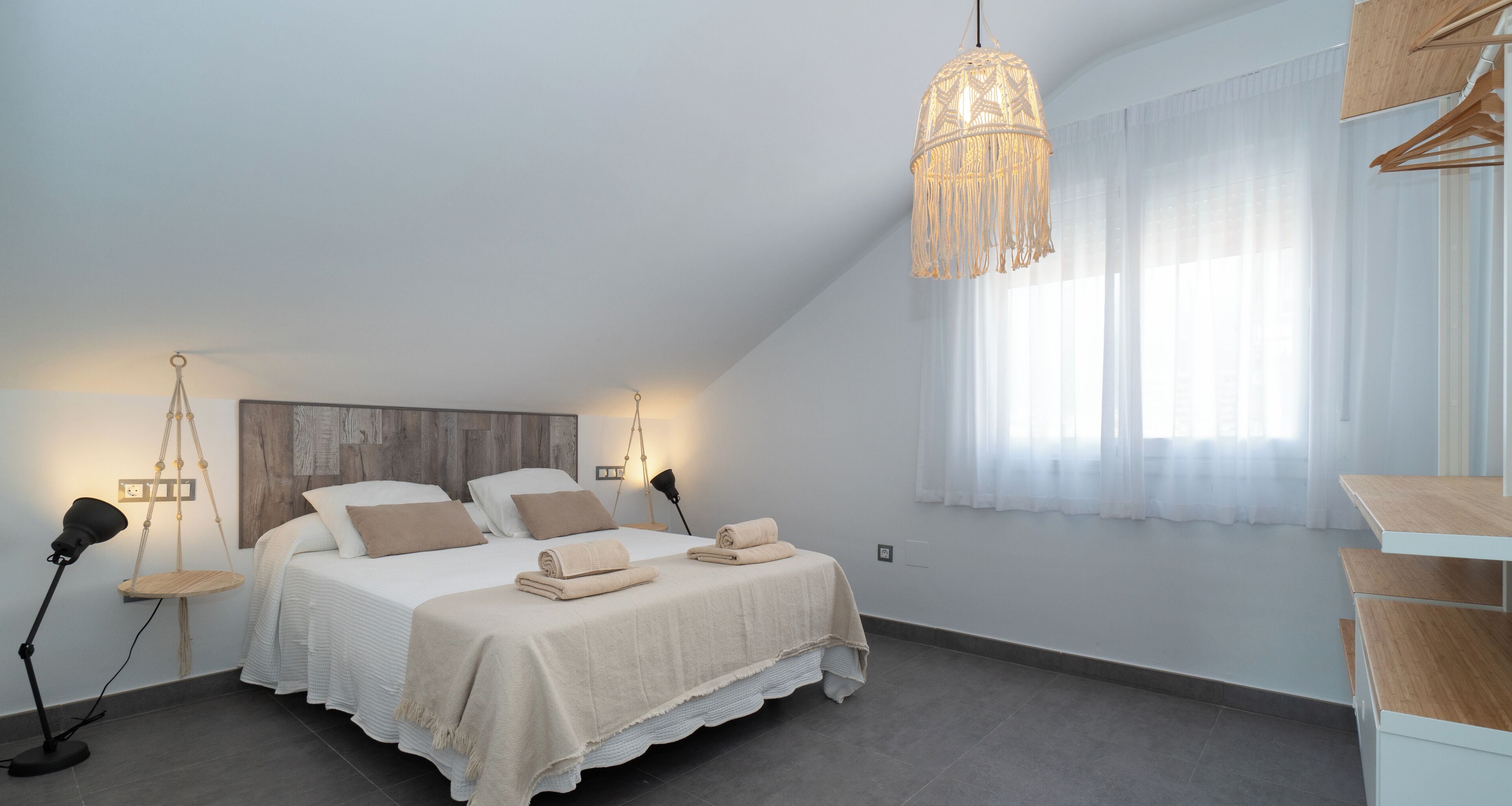 Un dormitorio con cama, lámparas de noche, lámpara colgante y ventana con cortinas translúcidas.