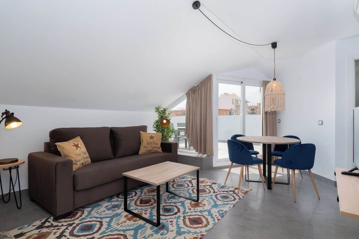 A modern living room with a brown sofa, a small wooden table, a patterned rug, a dining area with blue chairs, and a large window with curtains.