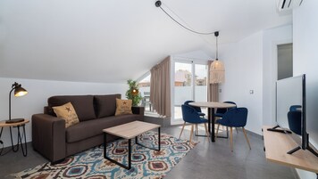 A modern living room with a brown sofa, a small wooden table, a patterned rug, a dining area with blue chairs, and a large window with curtains.
