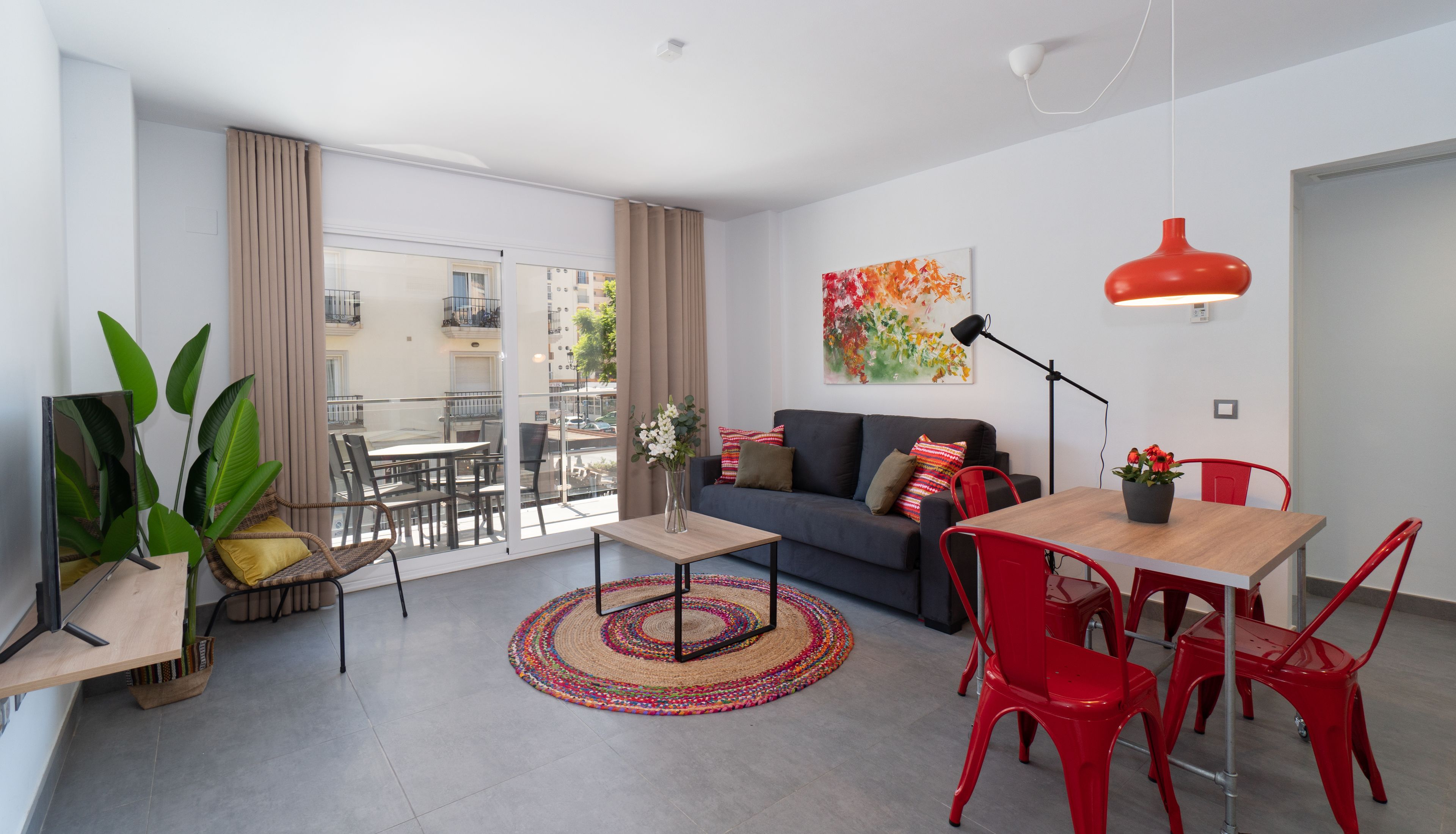 A modern living room with a sofa, a dining table, red chairs, a coffee table, a floor lamp, and a large window with a view of buildings.