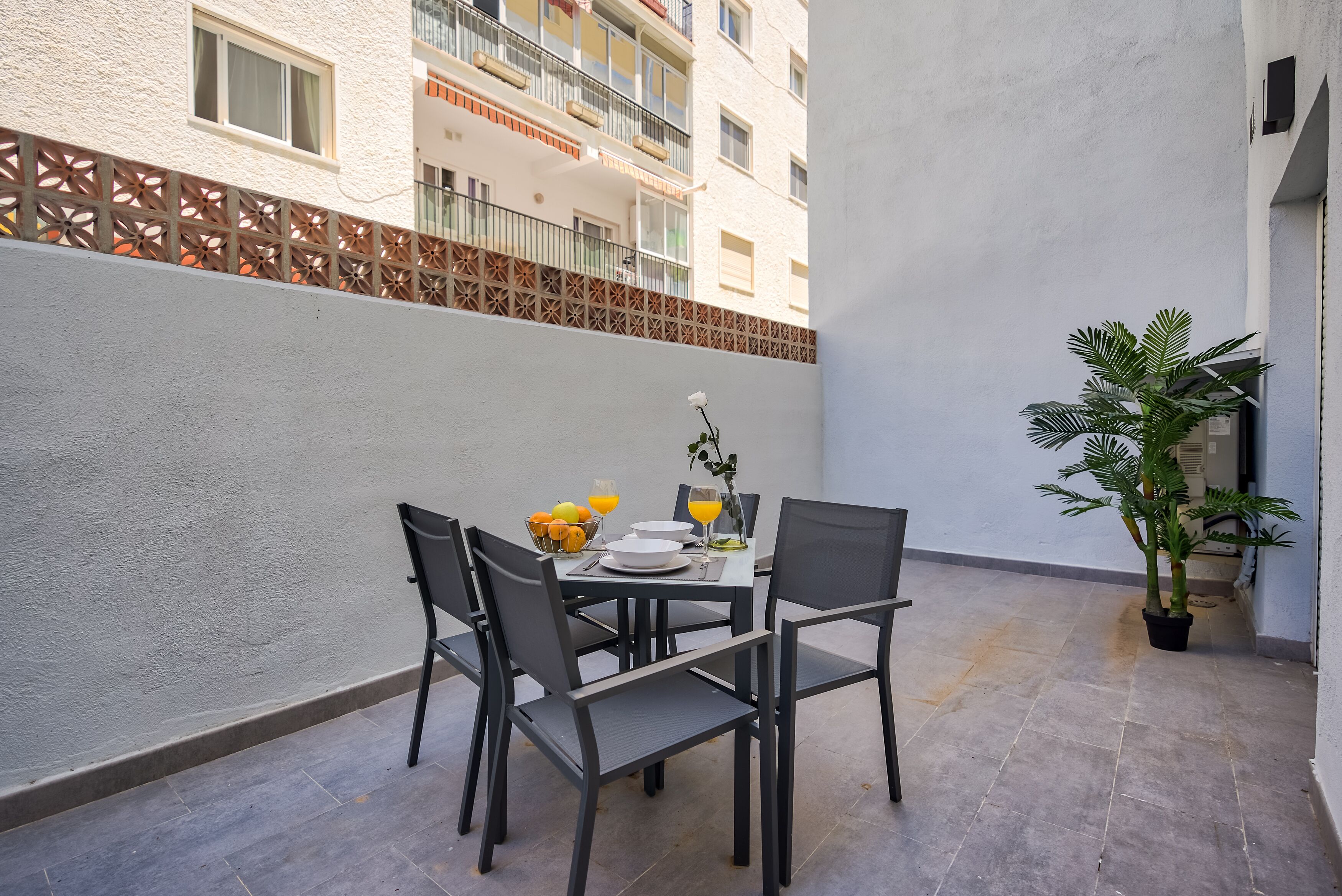 Un pequeño espacio de comedor al aire libre con una mesa, sillas, una planta en maceta y vistas a un edificio al fondo.