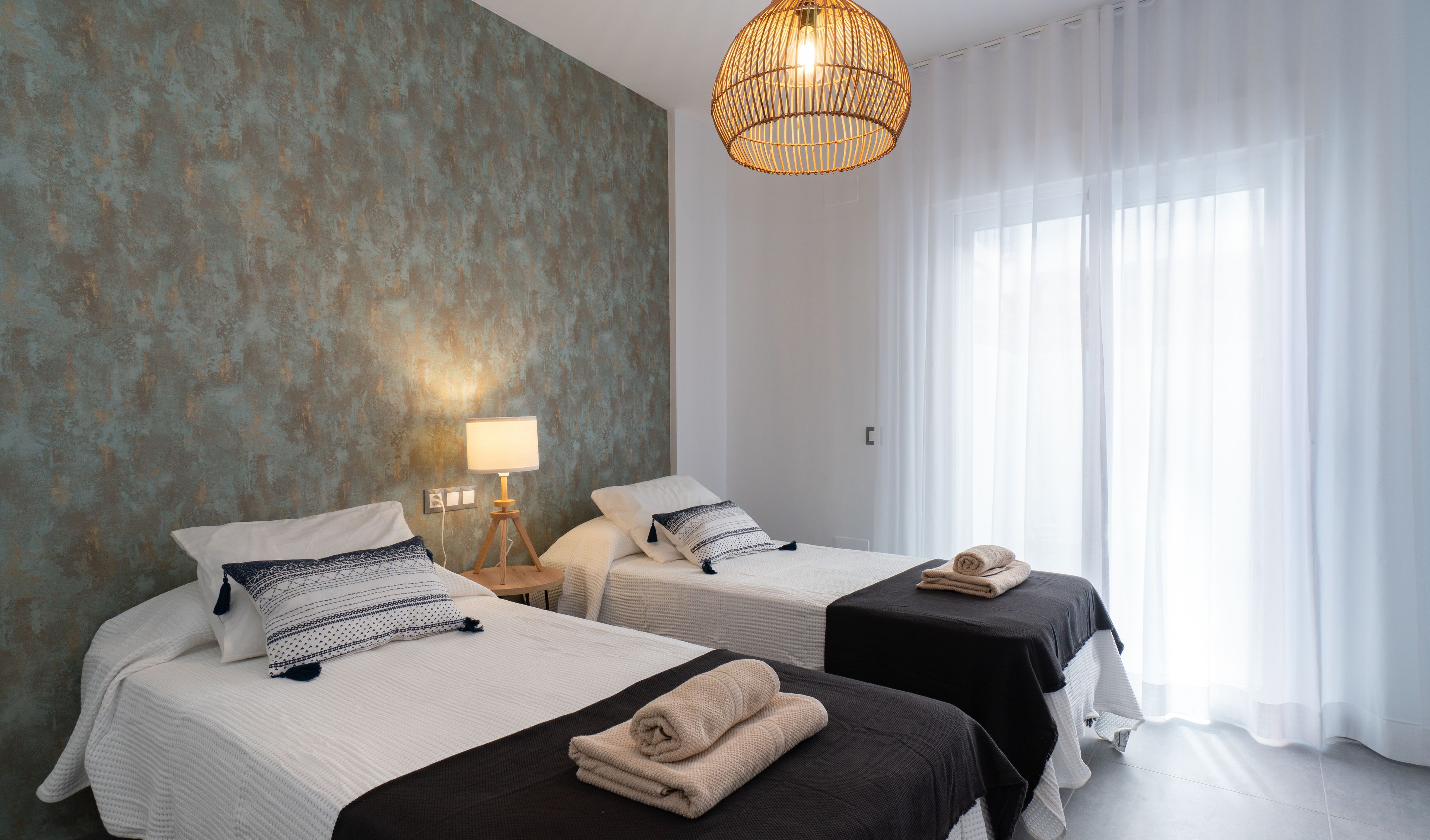 A hotel room with two beds, a lamp, a hanging light fixture, and sheer curtains.