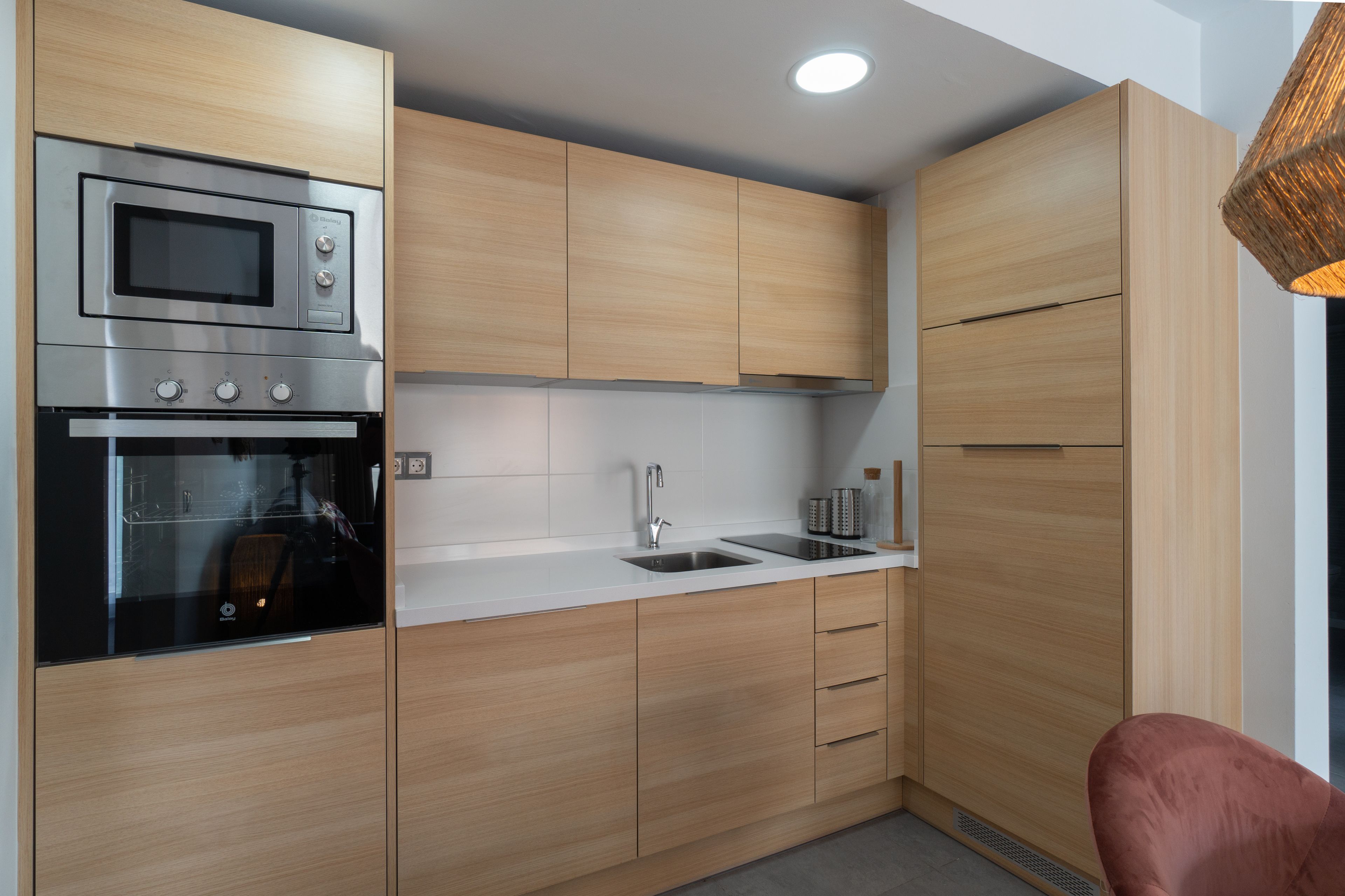 Une cuisine compacte dotée d’un micro-ondes, d’un four et d’un évier encastrés, de meubles en bois et d’un luminaire suspendu moderne.
