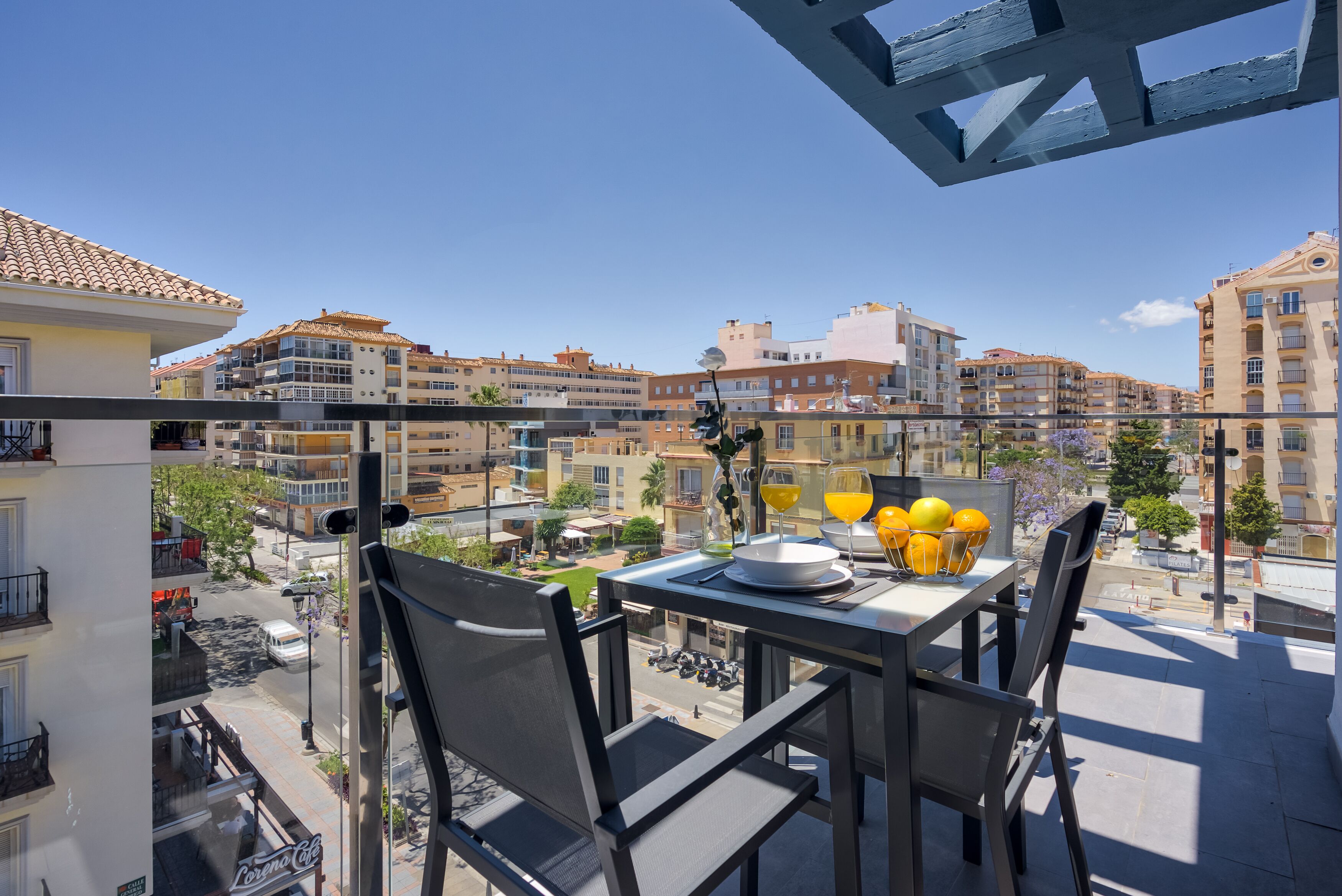 A balcony with a table set for breakfast, overlooking a street with buildings and parked cars.