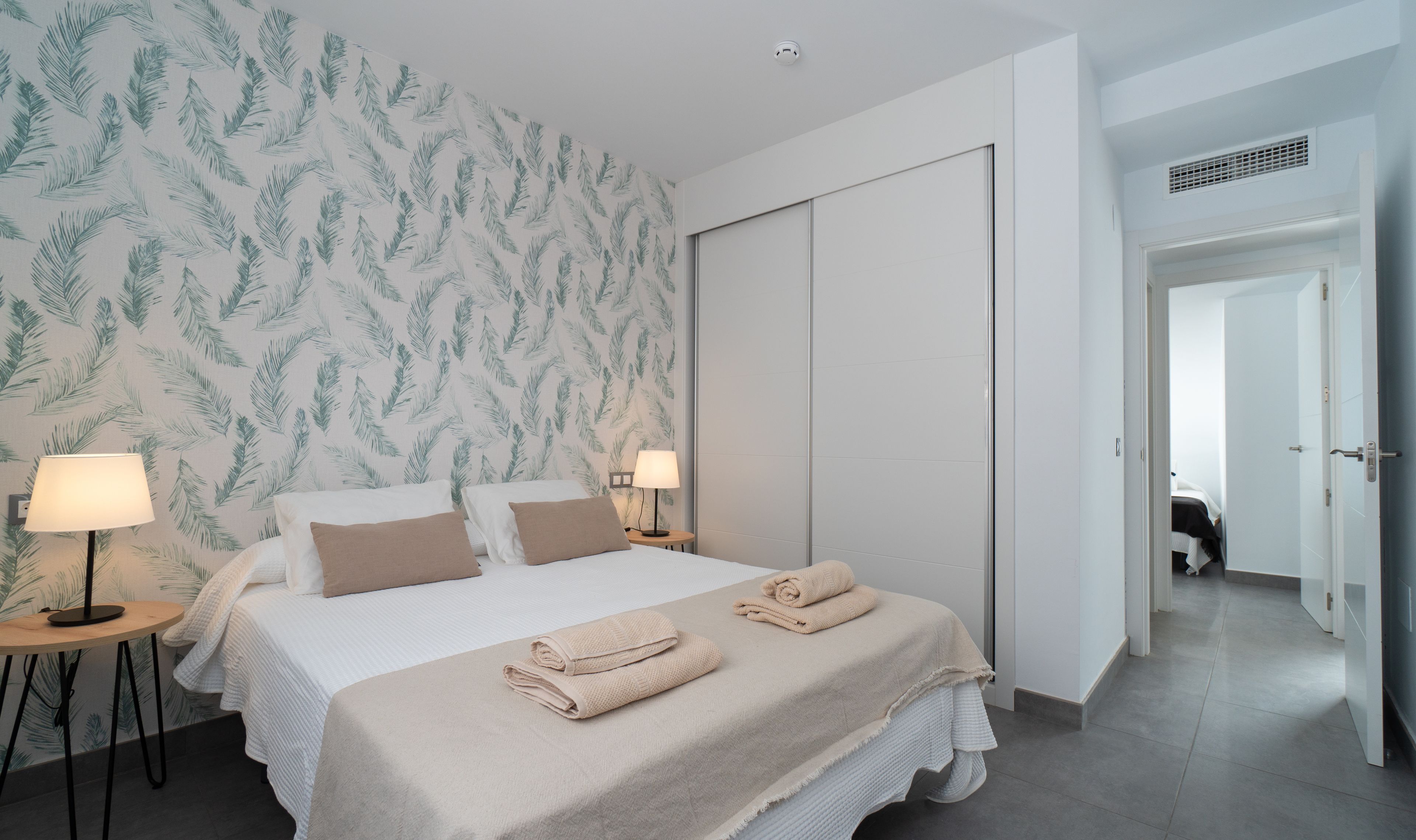 A modern bedroom with a large bed, two bedside lamps, a wardrobe, and a bathroom visible through an open door.