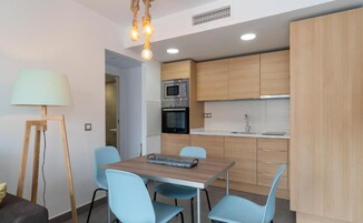 A compact kitchen with wooden cabinets, a microwave, an oven, and a sink. There is a dining table with chairs and a lamp.