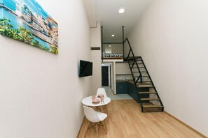 Quarto pequeno com parede branca, uma mesa redonda com cadeiras, um quadro e uma escada com corrimão preto.