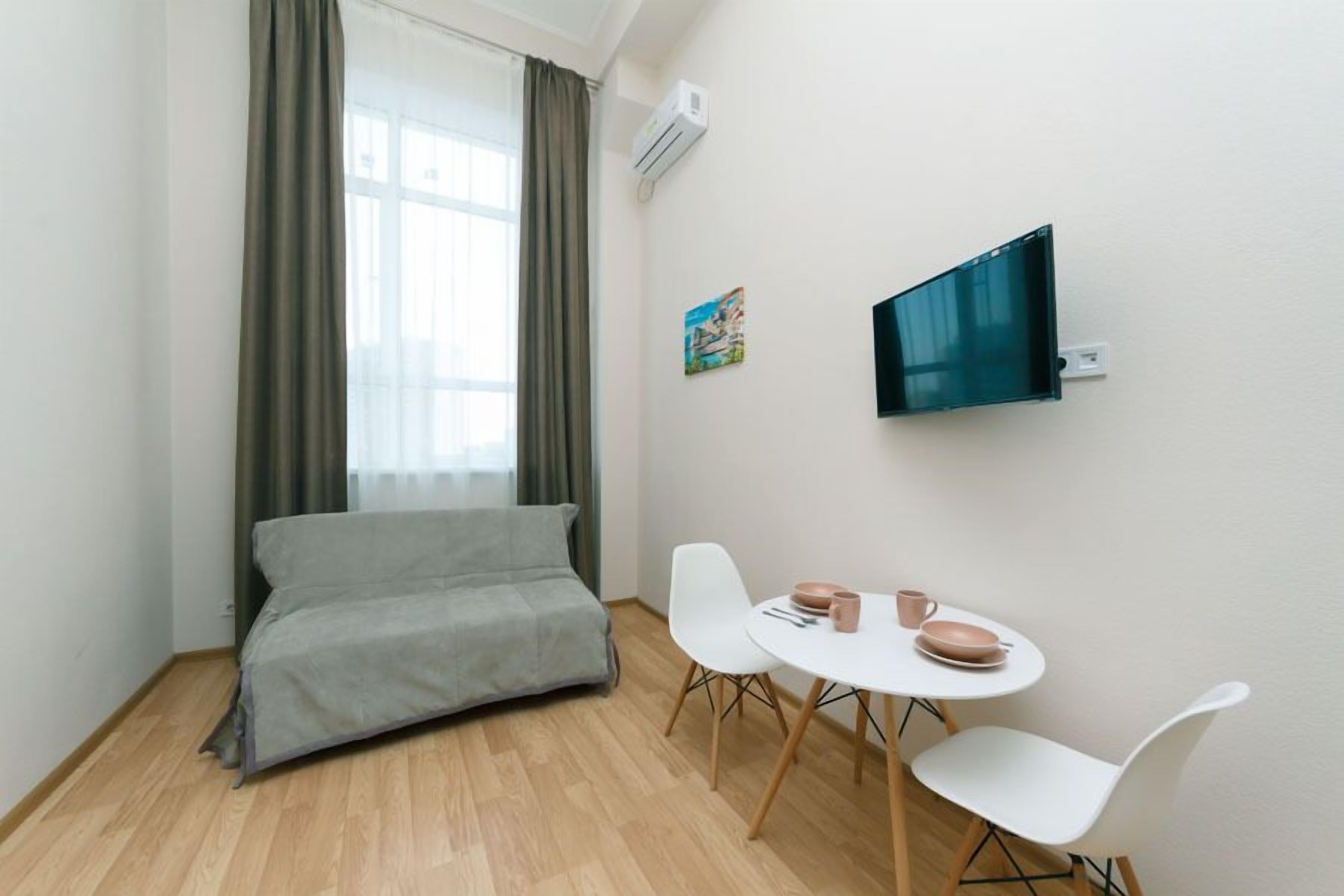 A small, modern room with a sofa, a dining table with chairs, a television mounted on the wall, and a window with curtains.