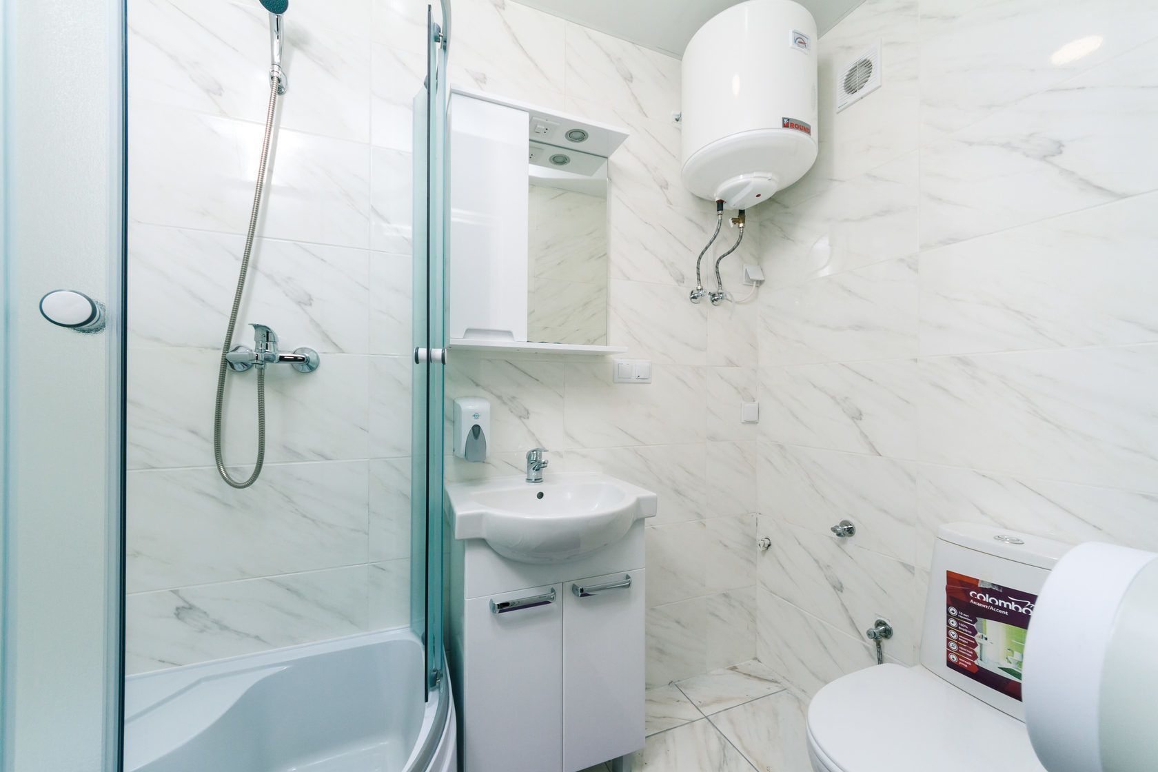 Un baño moderno con cabina de ducha de vidrio, un lavamanos blanco, un inodoro y un calentador de agua instalado.