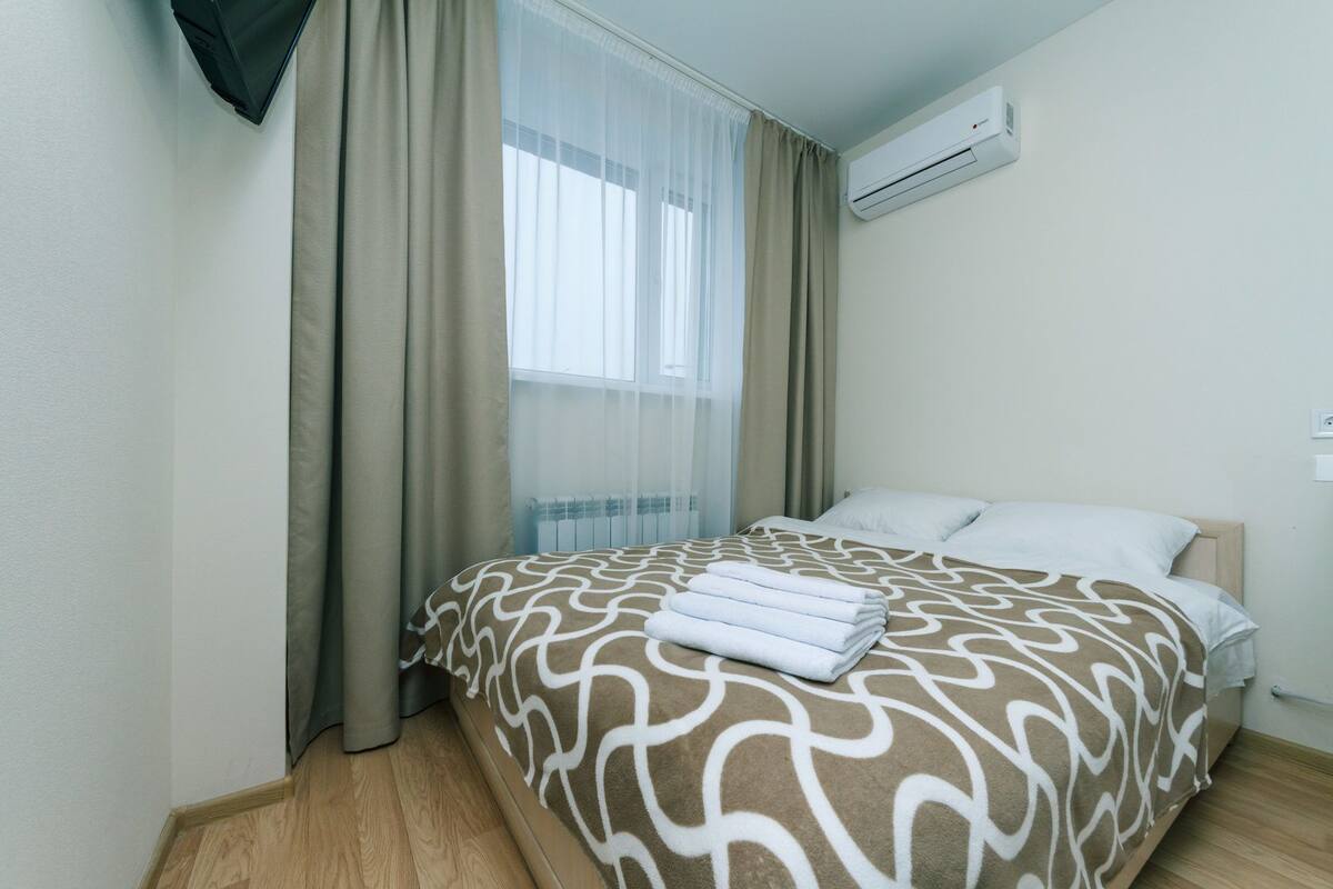 A hotel room with a bed, beige curtains, an air conditioning unit, and a window with sheer curtains.