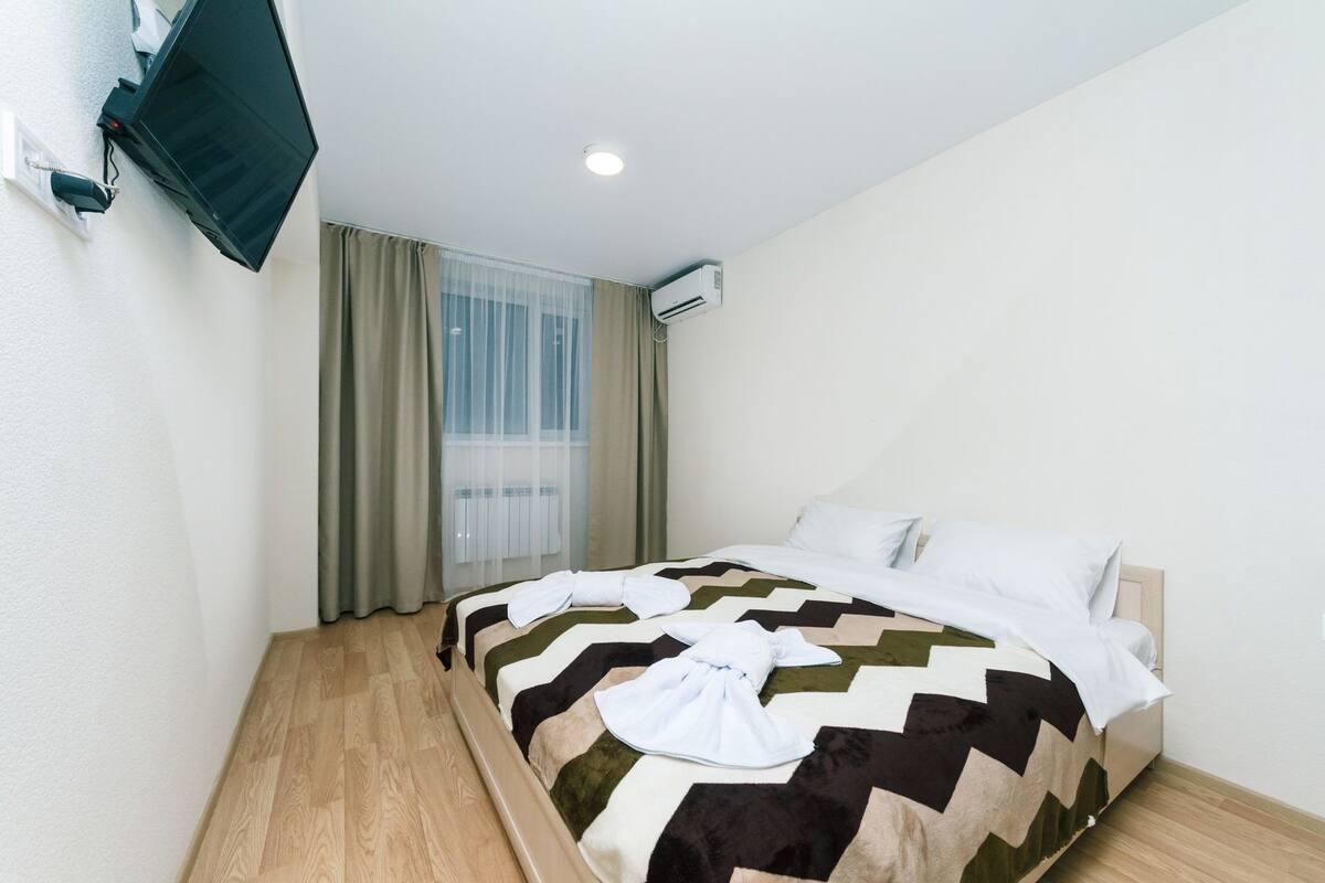 A hotel room with a bed, a TV mounted on the wall, an air conditioning unit, and a window with curtains.