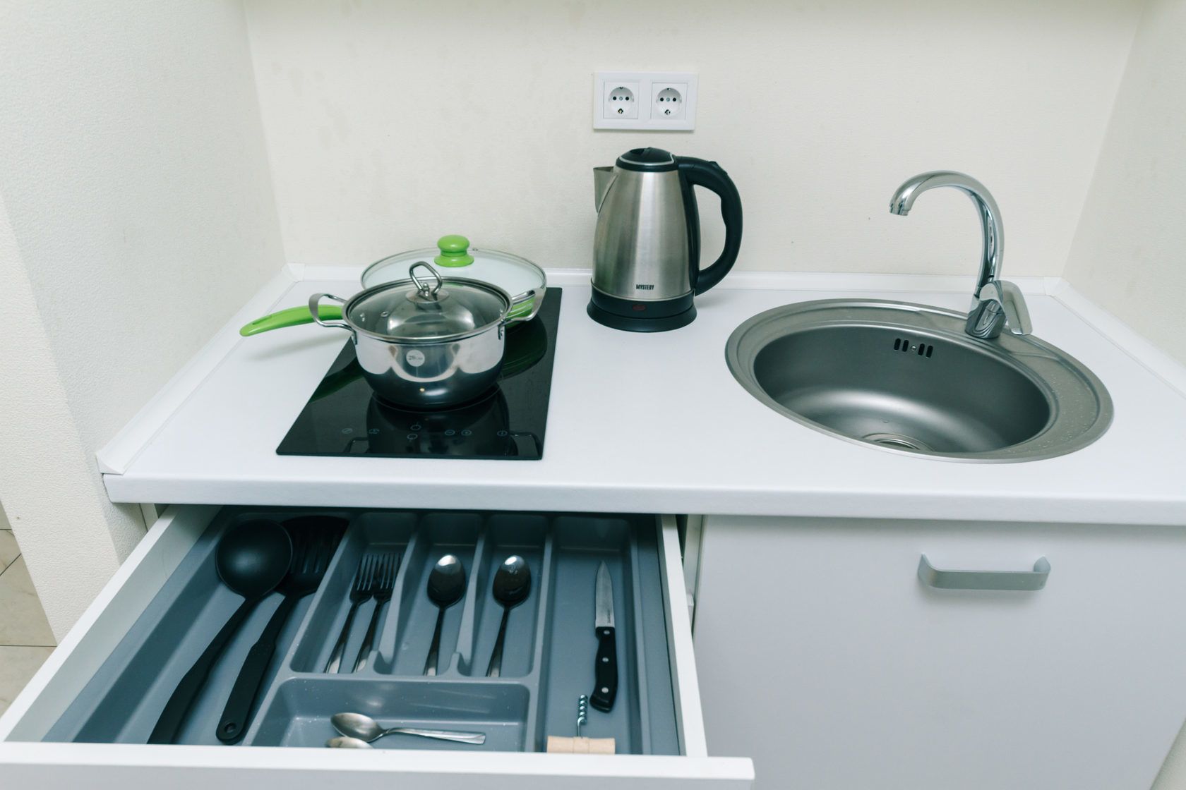 A compact kitchen with a stove, kettle, sink, and drawer containing utensils.