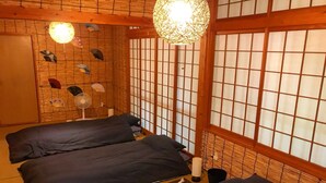 Una habitación japonesa tradicional con suelo de tatami, dos camas con ropa oscura, un abanico colgante, un celosía de papel y un aplique decorativo.