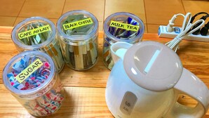 Cuatro frascos de vidrio etiquetados con diferentes ingredientes sobre una superficie de madera, una tetera eléctrica blanca y una regleta con cables.