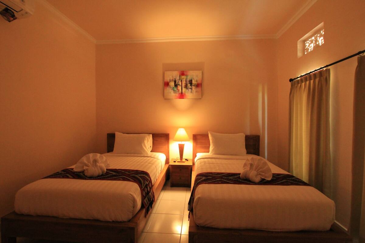 A hotel room with two beds, a nightstand with a lamp, a painting on the wall, and a window with curtains.