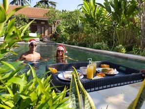 A poolside setting with two people in the water, a dining table with food and drinks, and lush greenery. - Ayuna Family Ubud (Ubud)