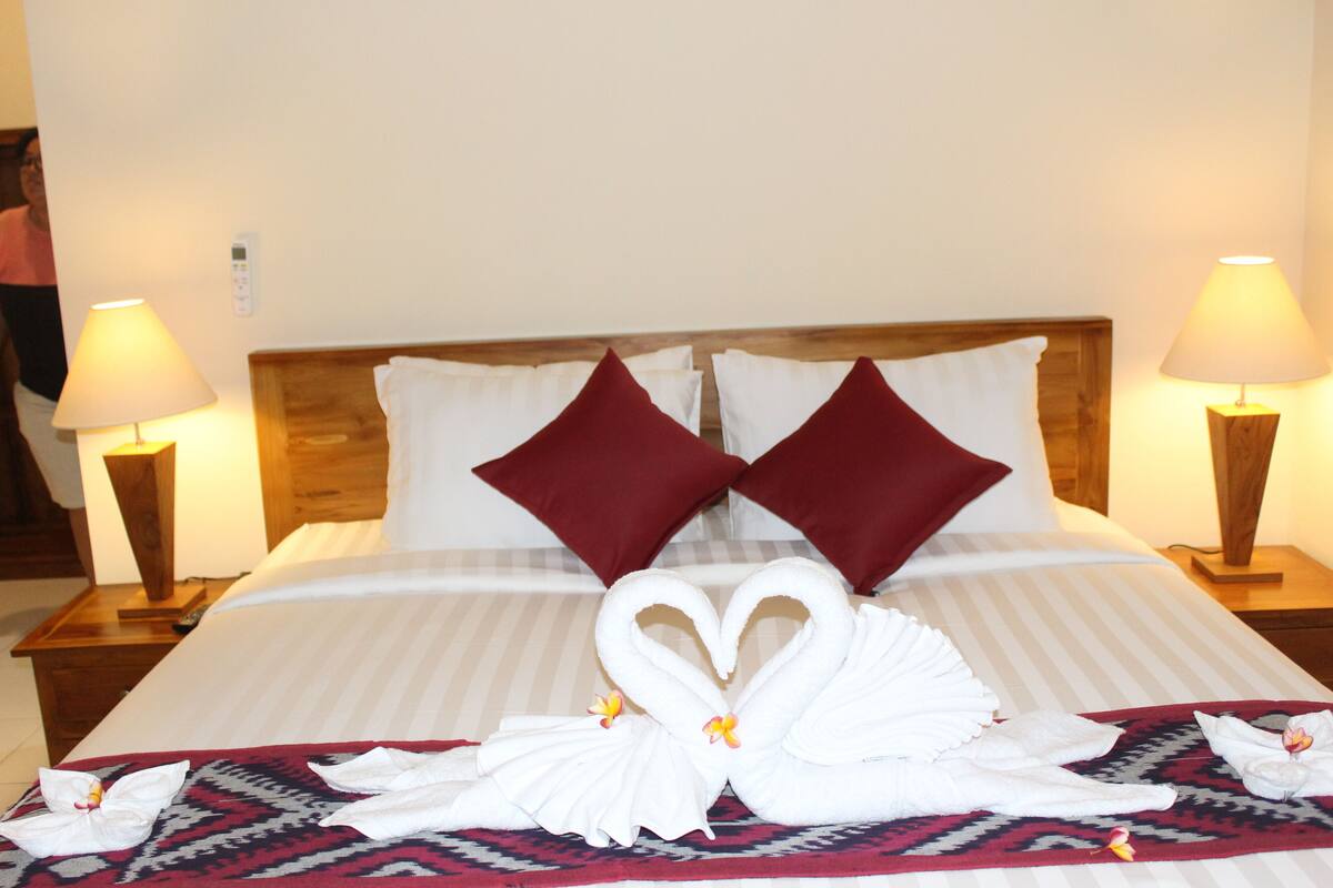 A hotel room with a bed, two red pillows, two bedside lamps, and a towel art arrangement on the bed.