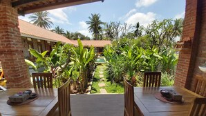 A covered patio with two wooden tables and chairs, surrounded by lush greenery and a view of a building with a tiled roof. - Ayuna Family Ubud (Ubud)