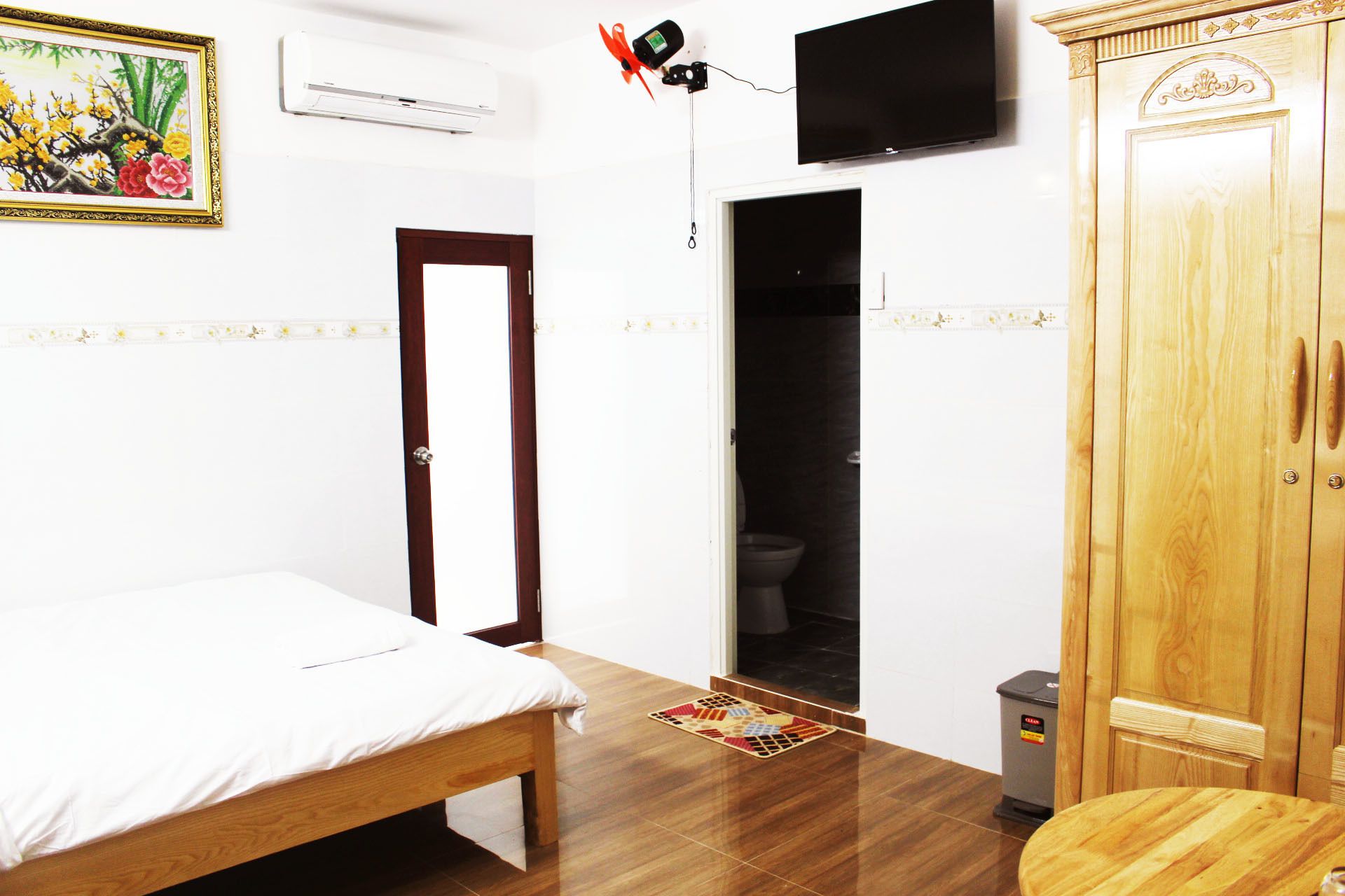 A room with a bed, a wooden wardrobe, a television mounted on the wall, and a small trash bin.