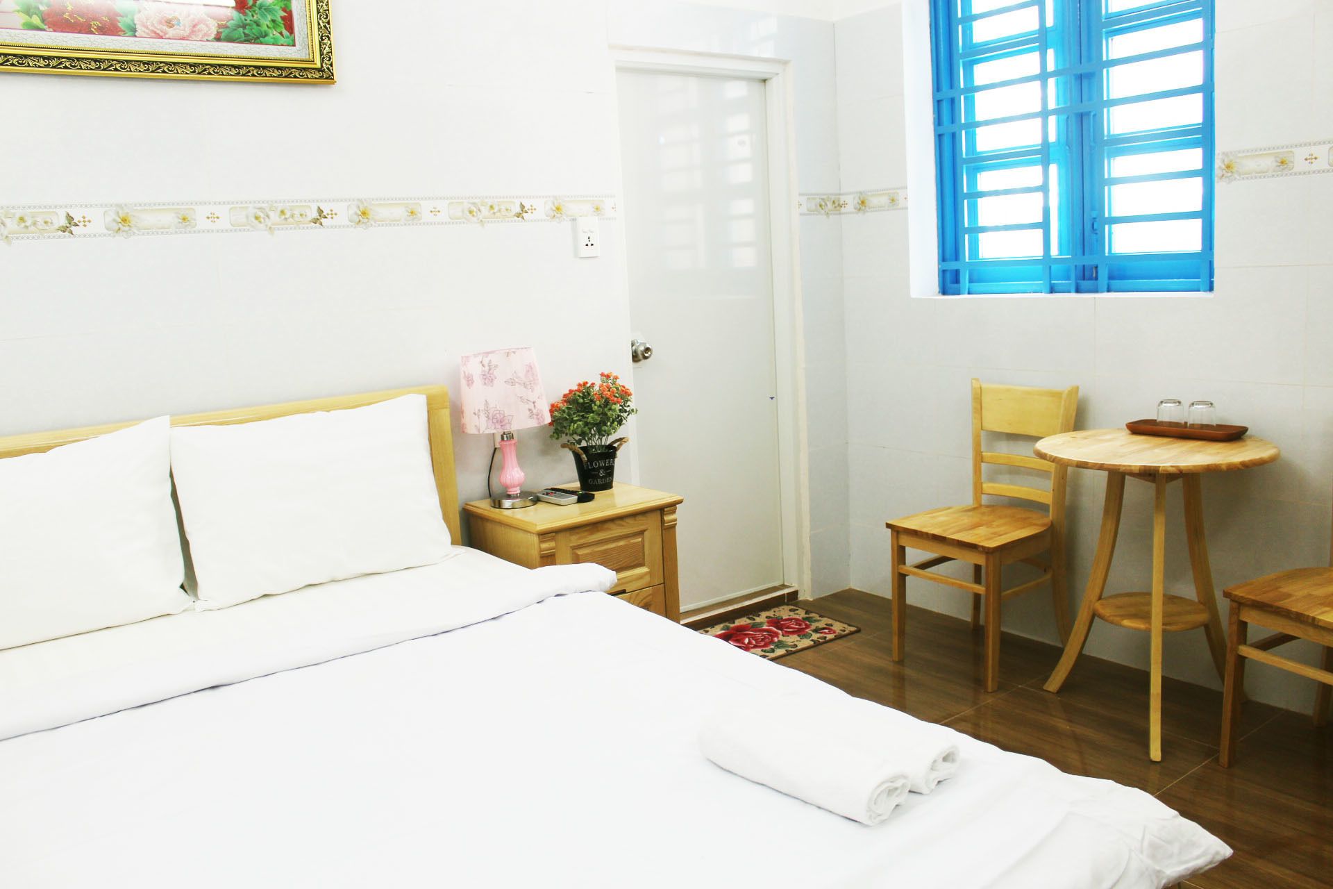 A hotel room with a bed, a small wooden table with a chair, a potted plant, and a window with blue shutters.