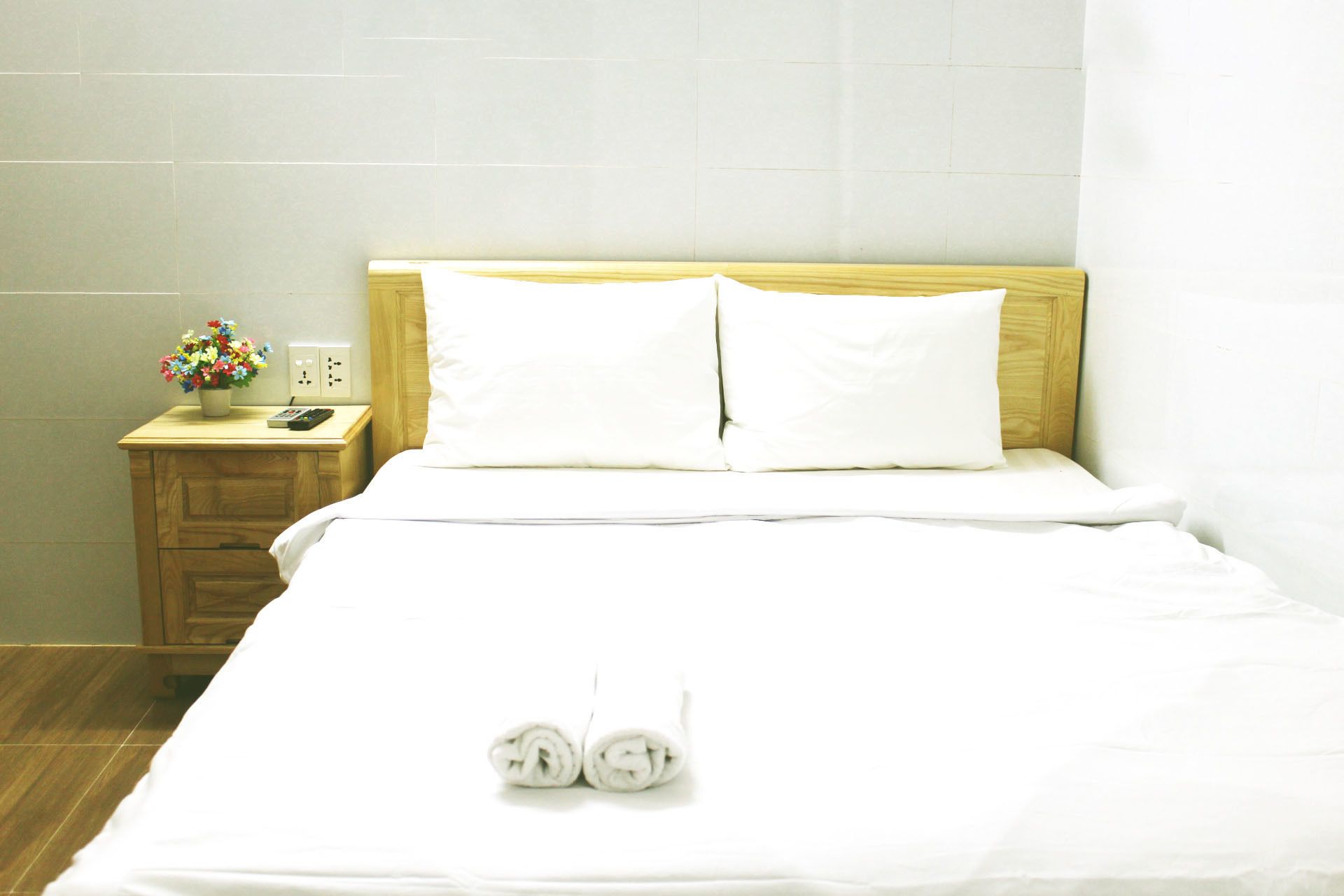 A hotel room with a wooden headboard, white bedding, a small bedside table with a phone and a flower vase, and a neatly folded towel on the bed.