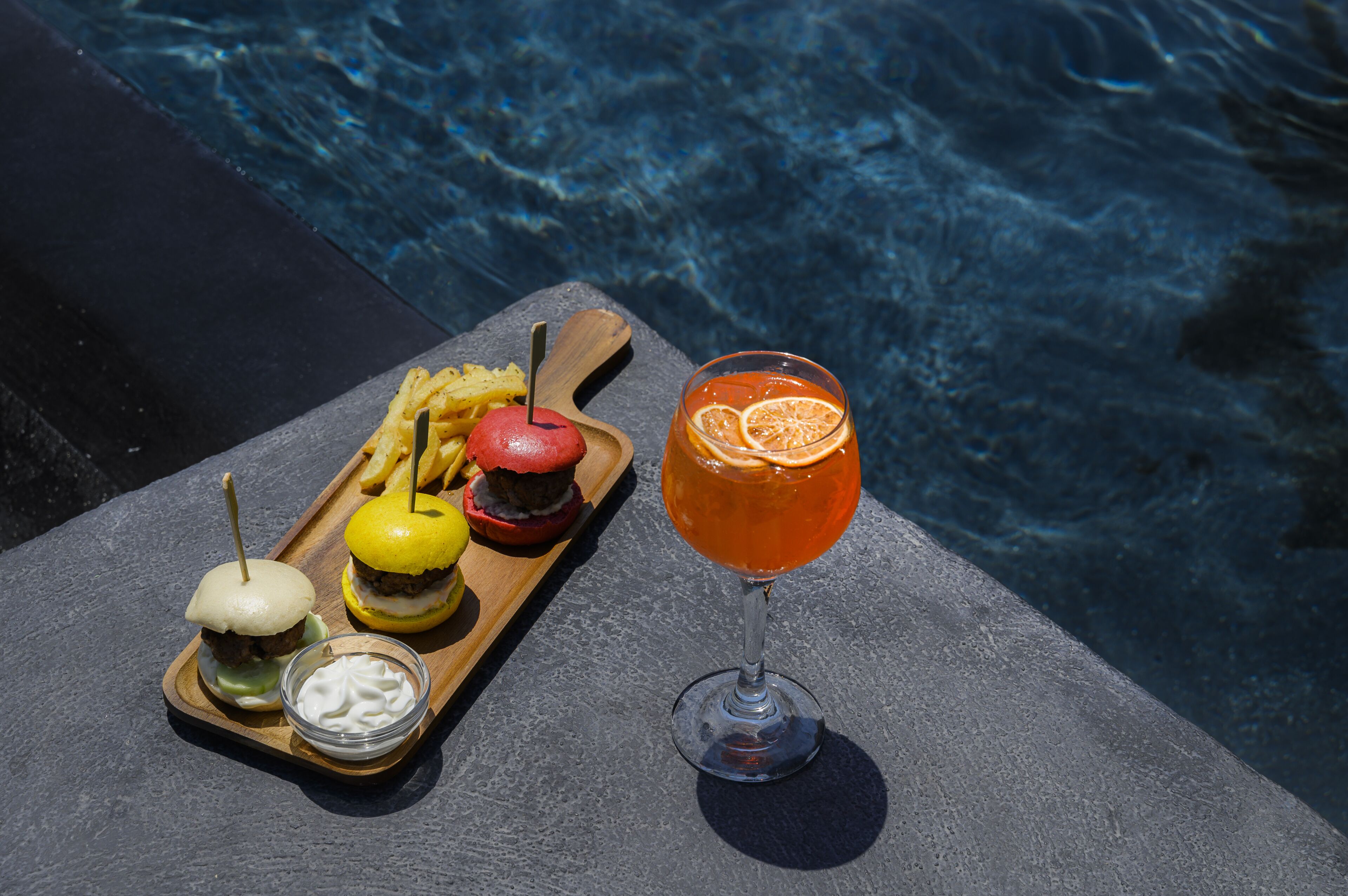 A wooden tray with a burger, a pickle, a lemon, and a glass of orange juice by a pool.