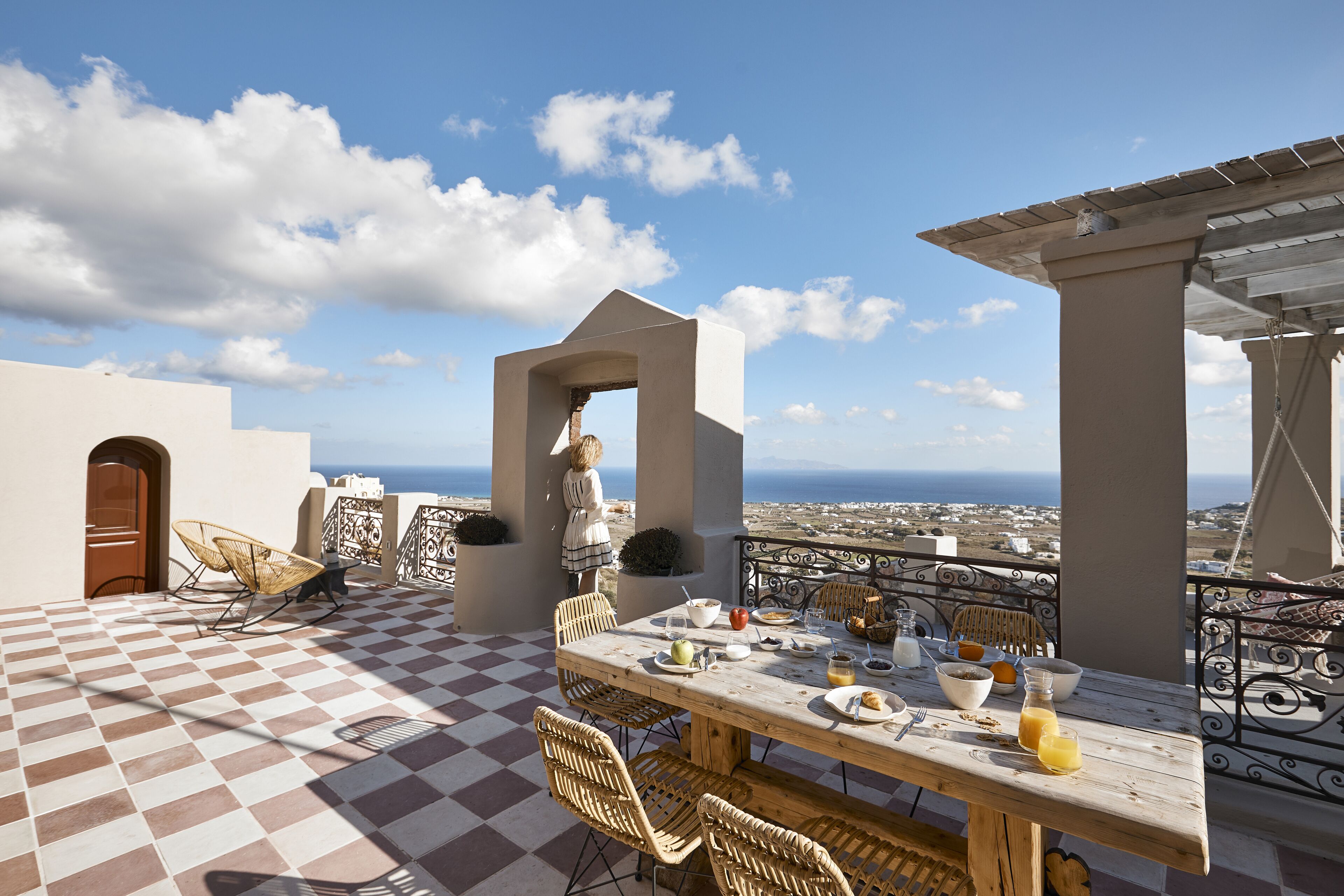 A terrace with a checkered floor, a wooden table set for breakfast, wicker chairs, and a view of the sea.