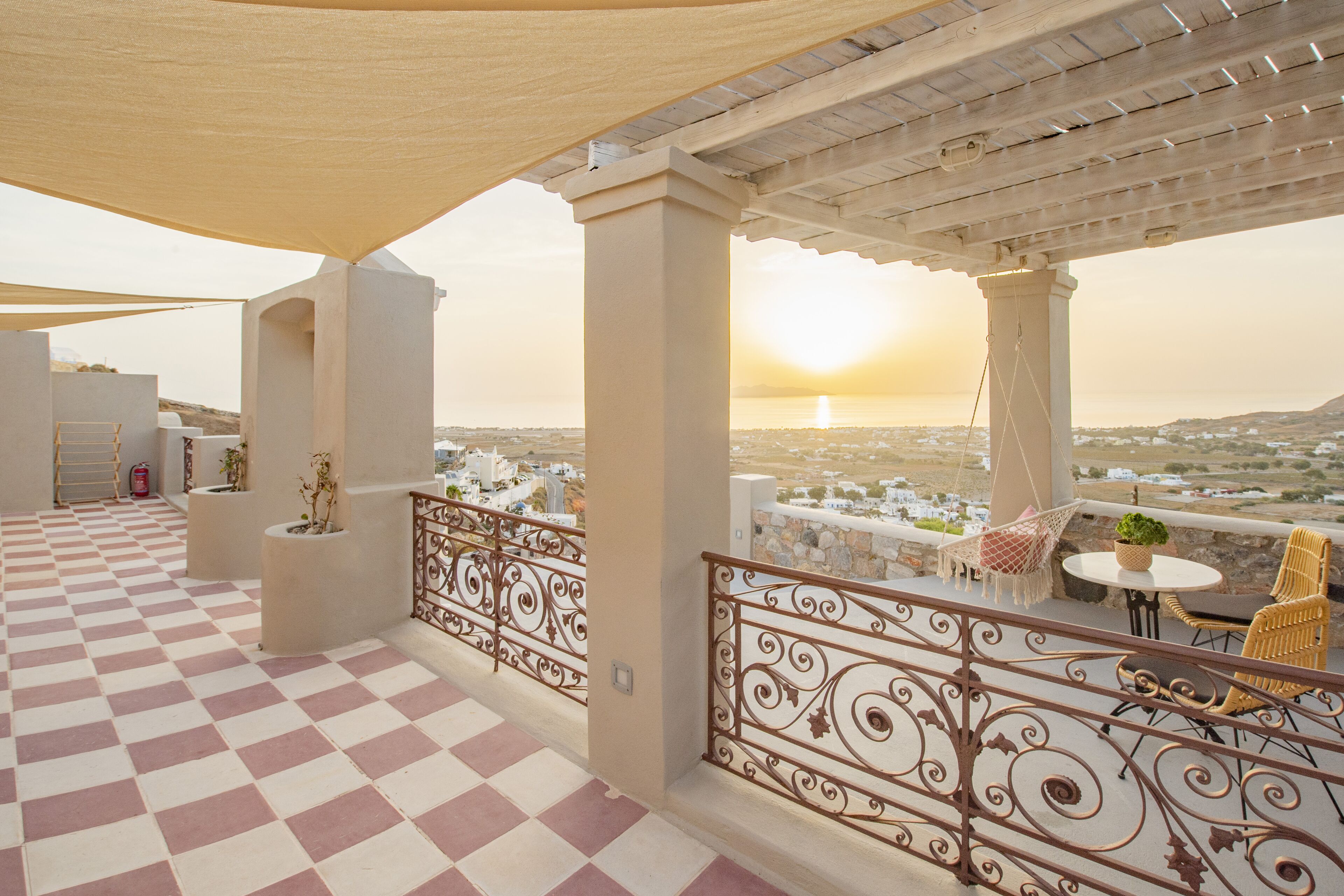 A terrace with a checkered floor, a pergola, and a view of the sunset over a cityscape.
