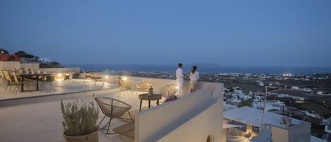 A rooftop terrace with outdoor seating, a potted plant, and a view of the city at dusk.