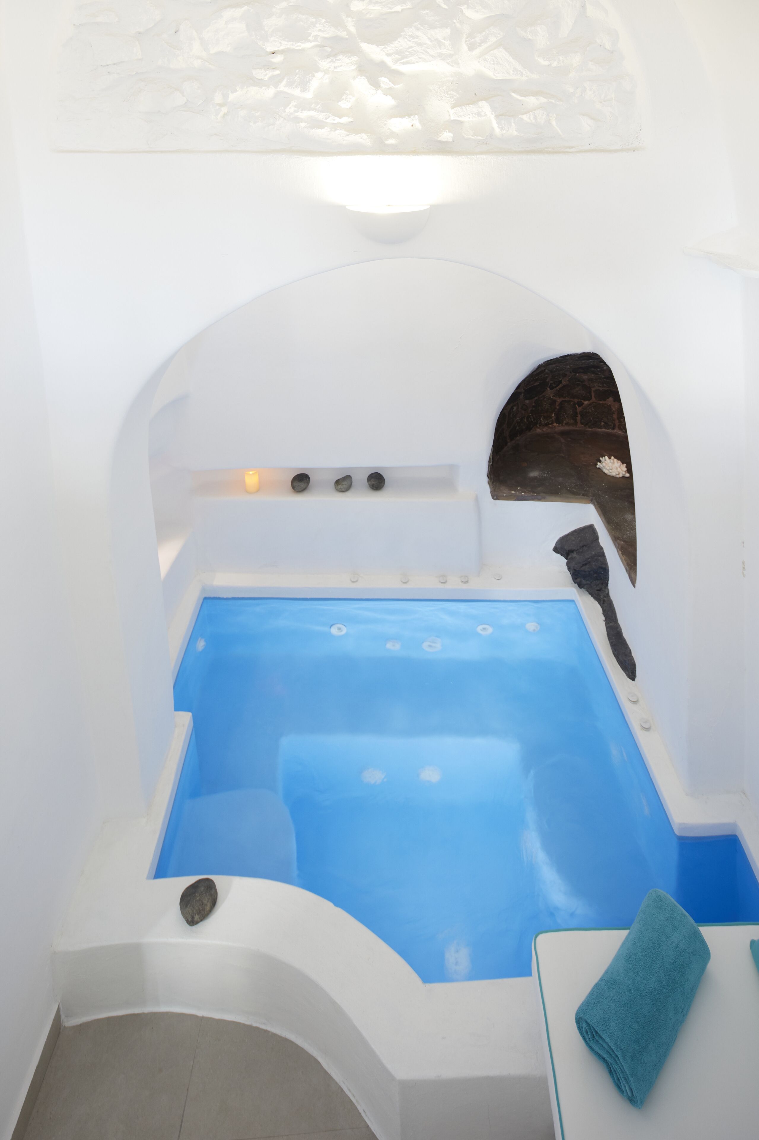 A white, arched bathtub with a blue interior, a stone wall, and a towel on a table.