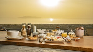 Una mesa de desayuno con diferentes alimentos y condimentos, frente a una vista panorámica del océano y una isla distante al atardecer.