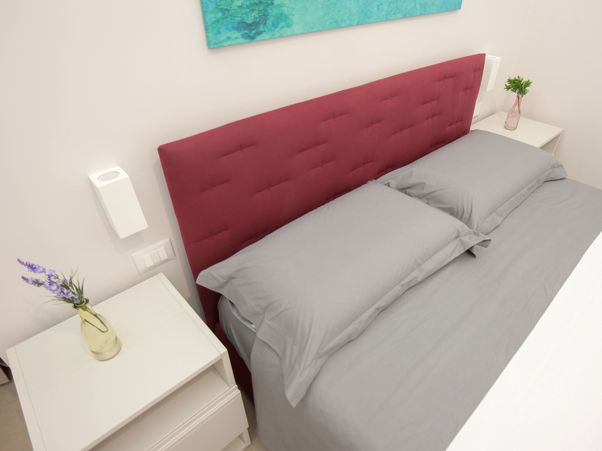 A neatly made bed with a red headboard, a white bedside table with a vase of flowers, and a wall-mounted light switch.