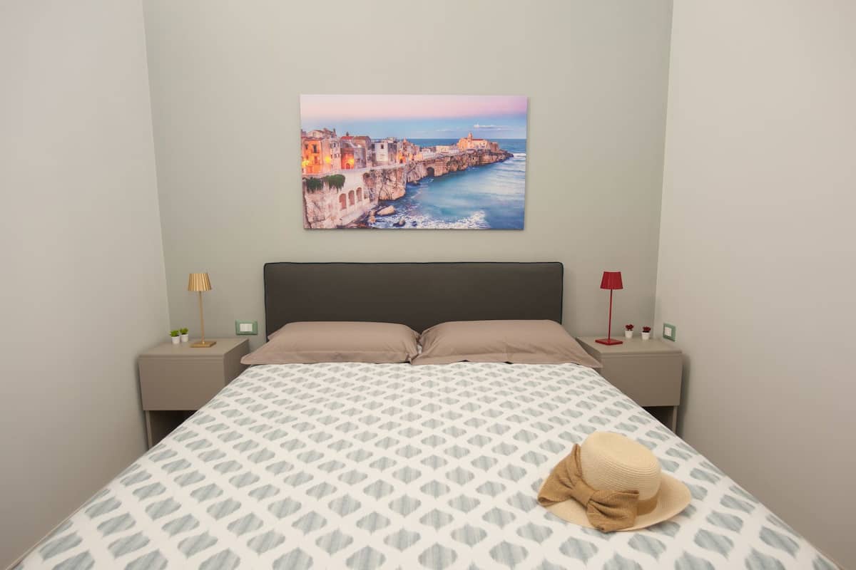 Quarto com cama, mesa de cabeceira, abajur, pintura de uma cidade costeira e um chapéu sobre a cama.