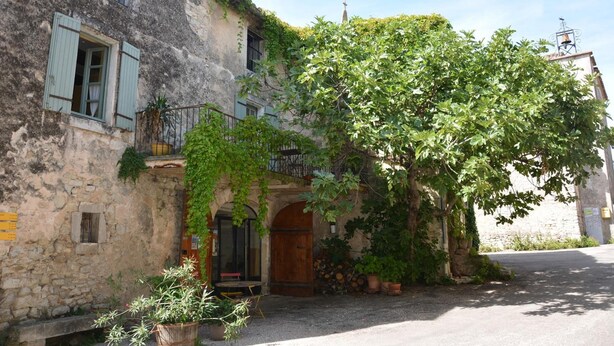 A rustic stone building with a wooden door, a balcony with metal railings, and a large tree in front.