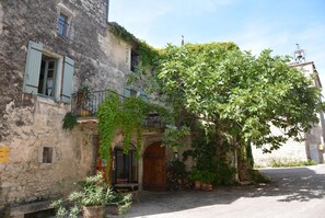 Un edificio rústico de piedra con puerta de madera, un balcón con barandilla de metal y un gran árbol en la parte delantera.