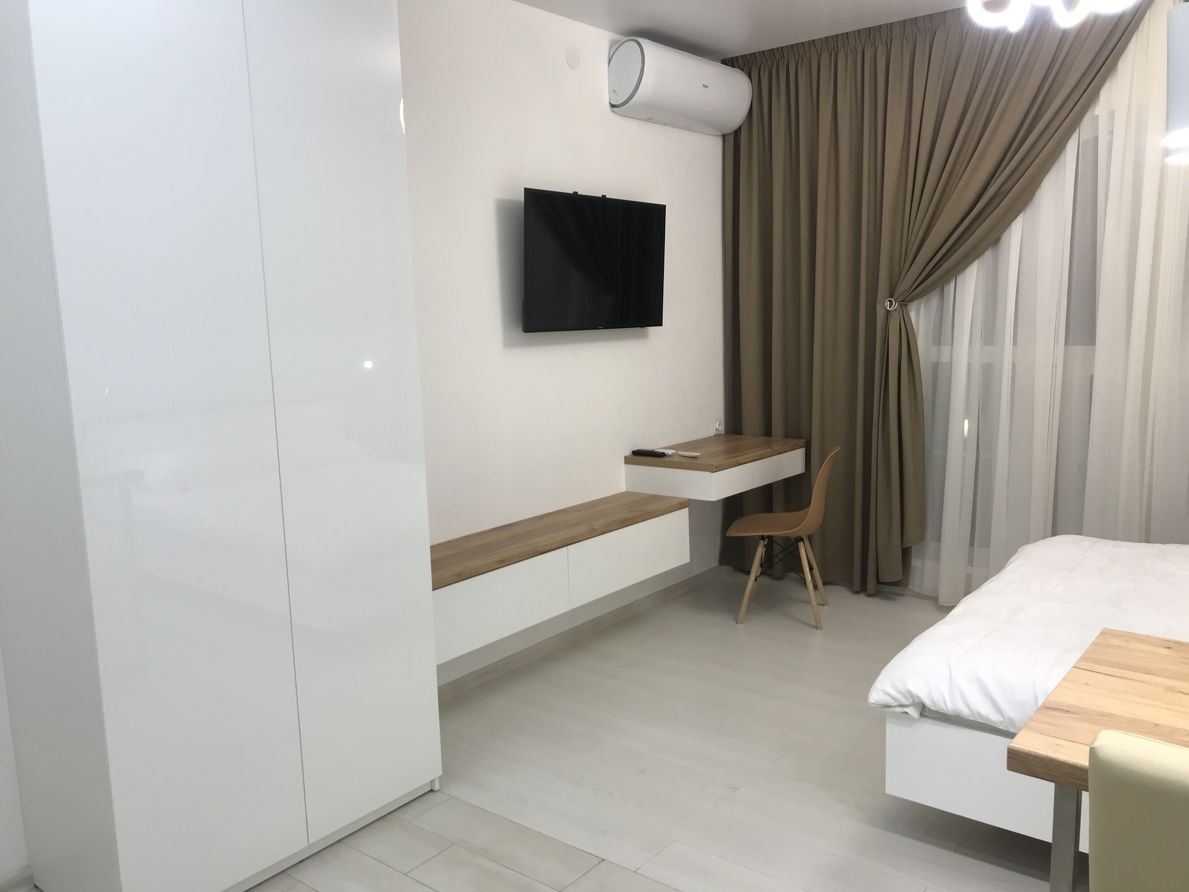 A modern hotel room with a bed, a desk with a chair, a mounted TV, and an air conditioning unit.