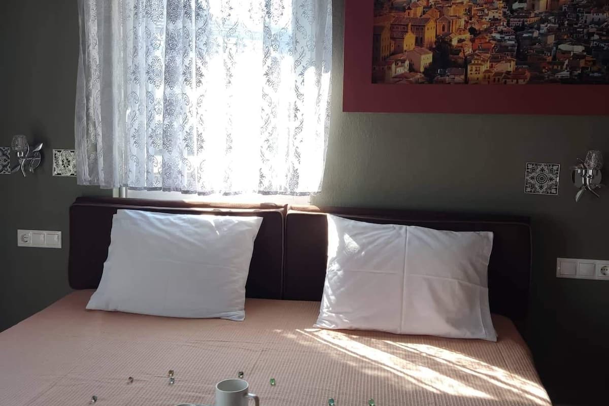 Cama bem arrumada com travesseiros brancos, bandeja com xícaras e pires, e um quadro de uma paisagem urbana na parede.