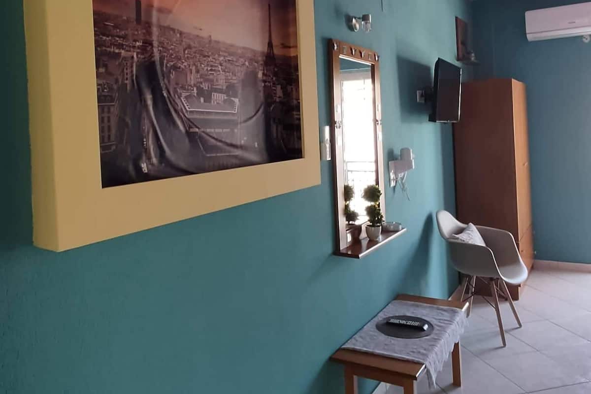 Quarto com paredes verde-água, uma grande pintura emoldurada de paisagem urbana, uma pequena mesa com um laptop, uma cadeira e um espelho.