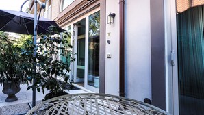 An outdoor seating area with a wicker table and umbrella, a potted plant, and a building entrance with glass doors and a doorbell. - Corte Merighi Charming Rooms & Breakfast (Verona)