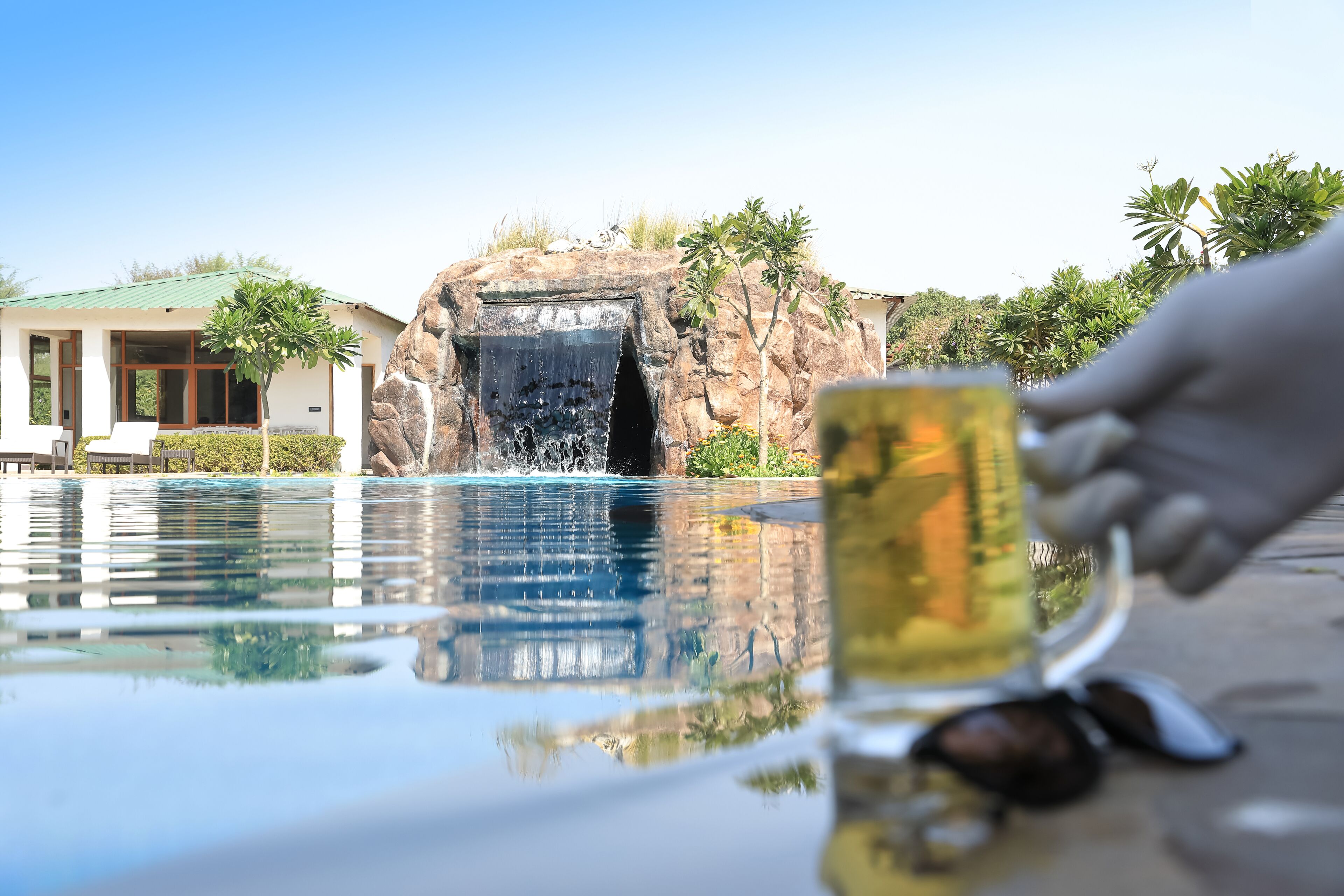 A pool with a waterfall feature, a glass of a beverage, and a hand holding the glass.