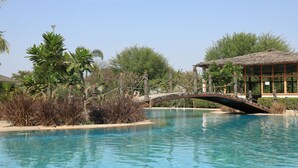 Una piscina con un puente, rodeada de vegetación y un edificio con techo de paja.