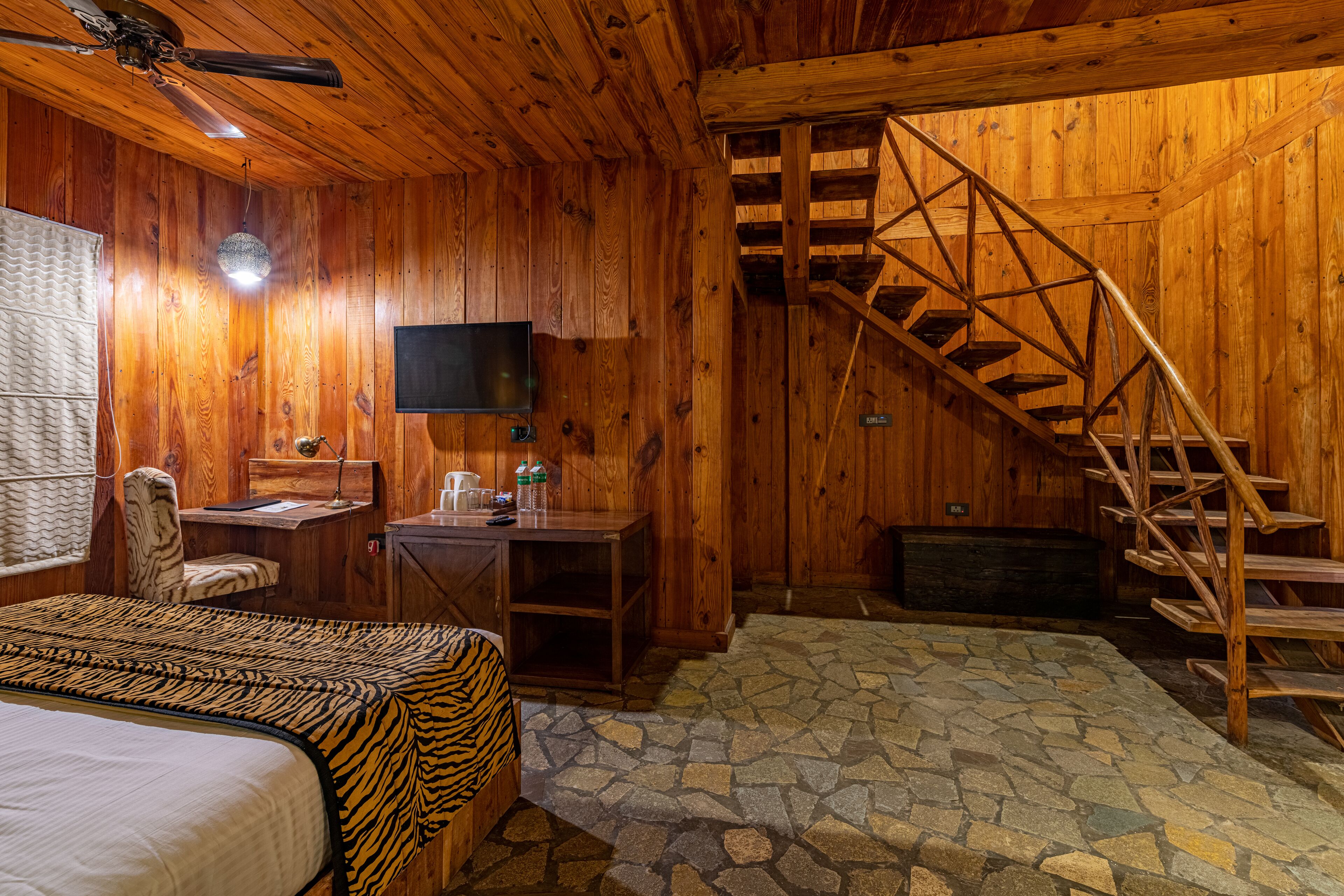 Ein Zimmer in einer Holzhütte mit Bett, Schreibtisch, Stuhl, Fernseher und einer Holztreppe.