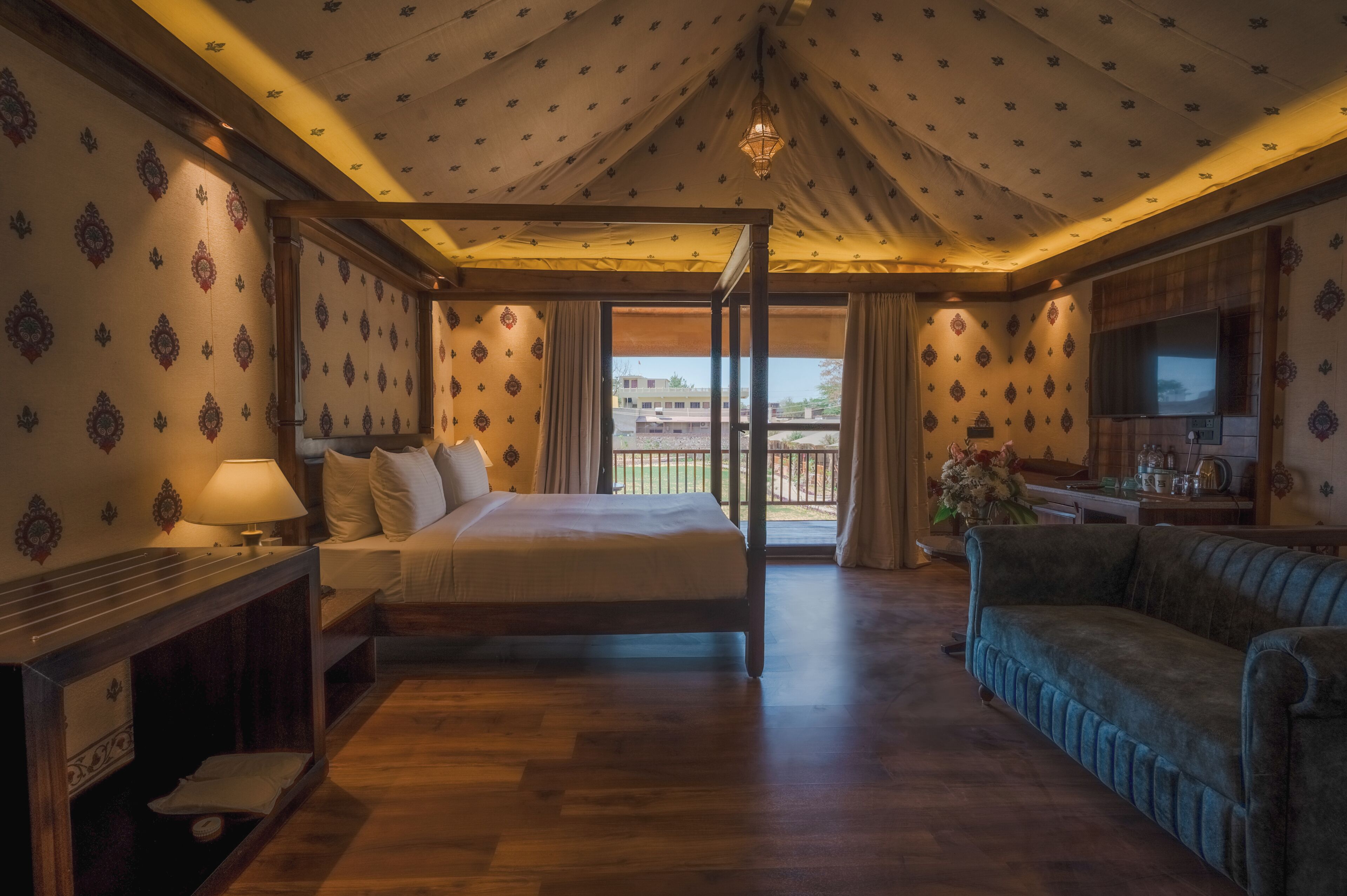 Habitación de hotel con cama, sofá, escritorio con lámpara, televisor y balcón con vistas.