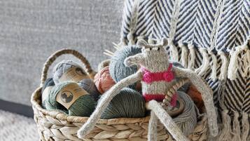 Ein Korbwägelchen voller Wollknäuel auf einem gemusterten Teppich, ein gestricktes Spielzeug und ein graues Sofa mit Decke im Hintergrund.