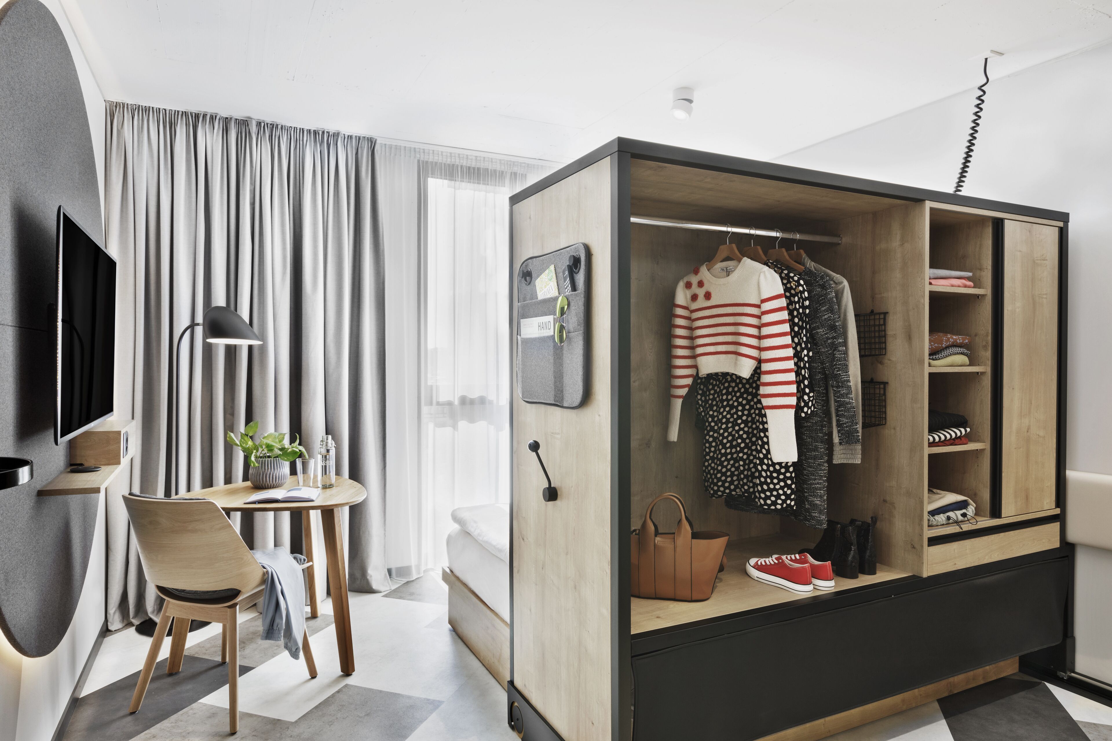 A modern hotel room with a wooden wardrobe, a desk with a chair, a TV, and a large window with curtains.