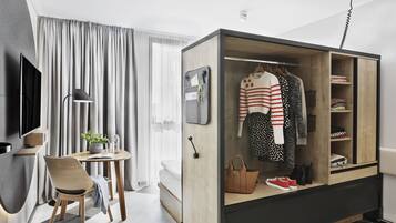 A modern hotel room with a wooden wardrobe, a desk with a chair, a TV, and a large window with curtains.
