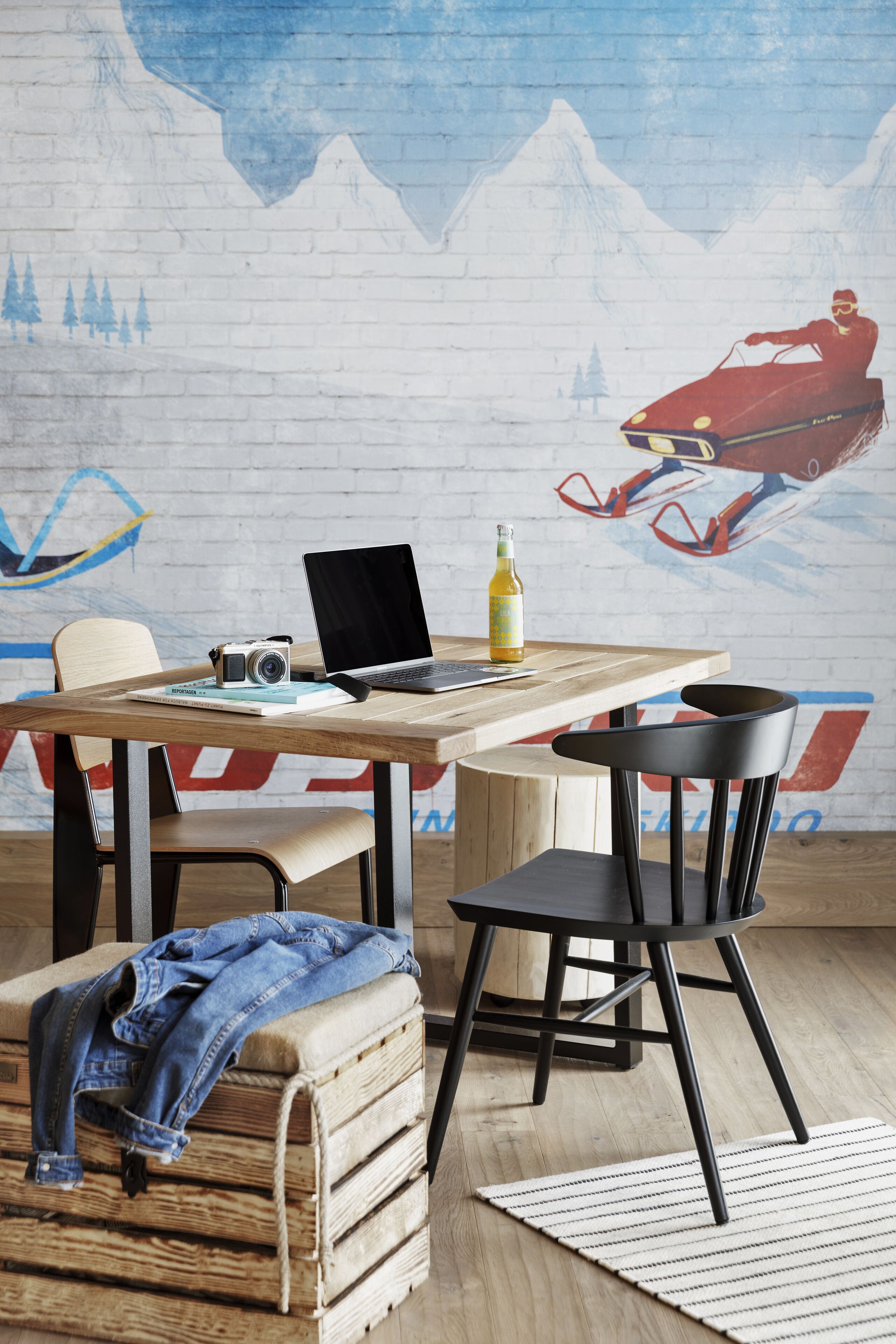 A room with a wooden table, a laptop, a camera, and a bottle. There is a chair with a denim jacket on it and a suitcase. The wall has a mural of a person on a motorcycle.