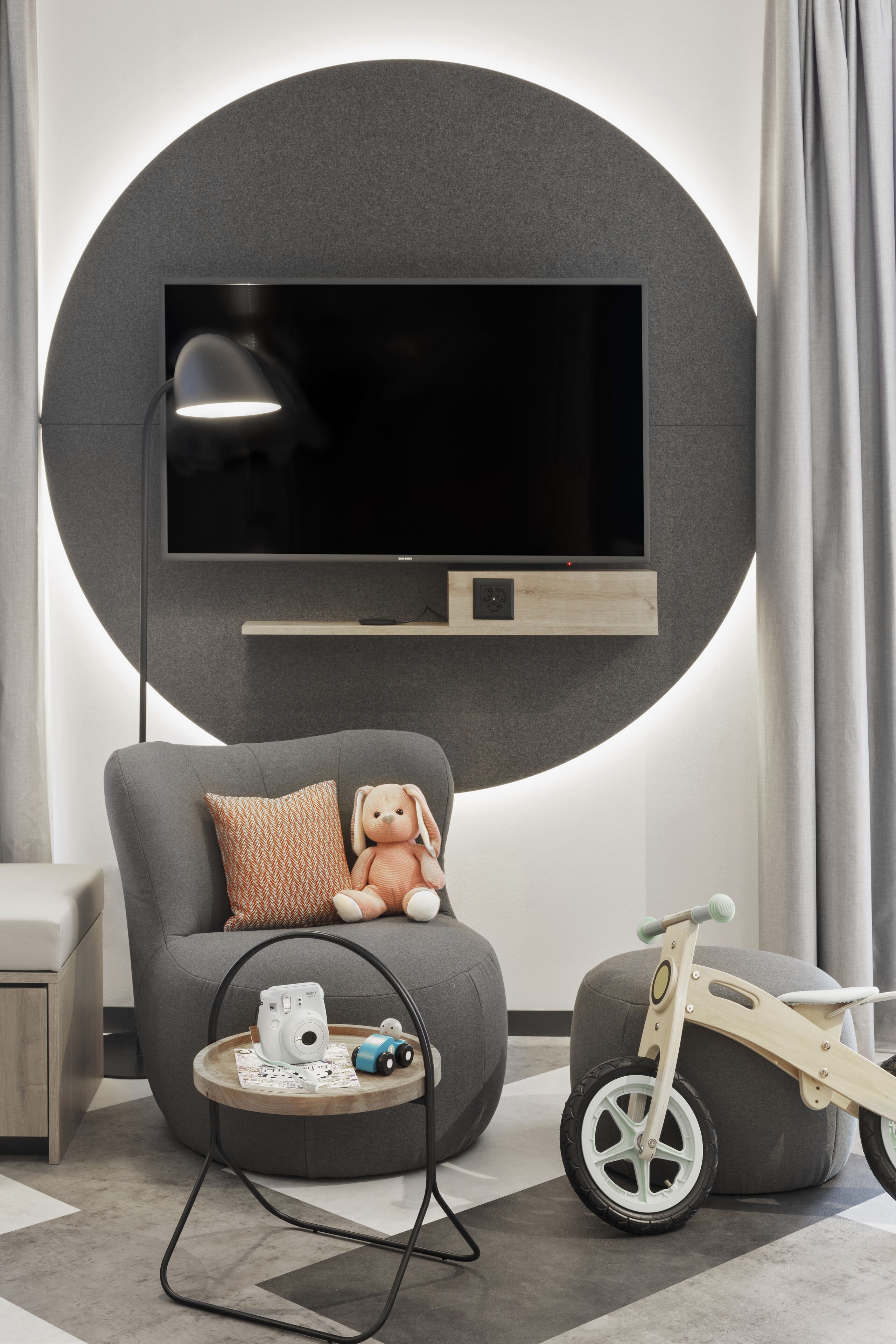 Un soggiorno moderno con una grande televisione a schermo piatto montata a parete, un divano grigio con un cuscino, un piccolo tavolino con una fotocamera e una macchinina, e una triciclo di legno.