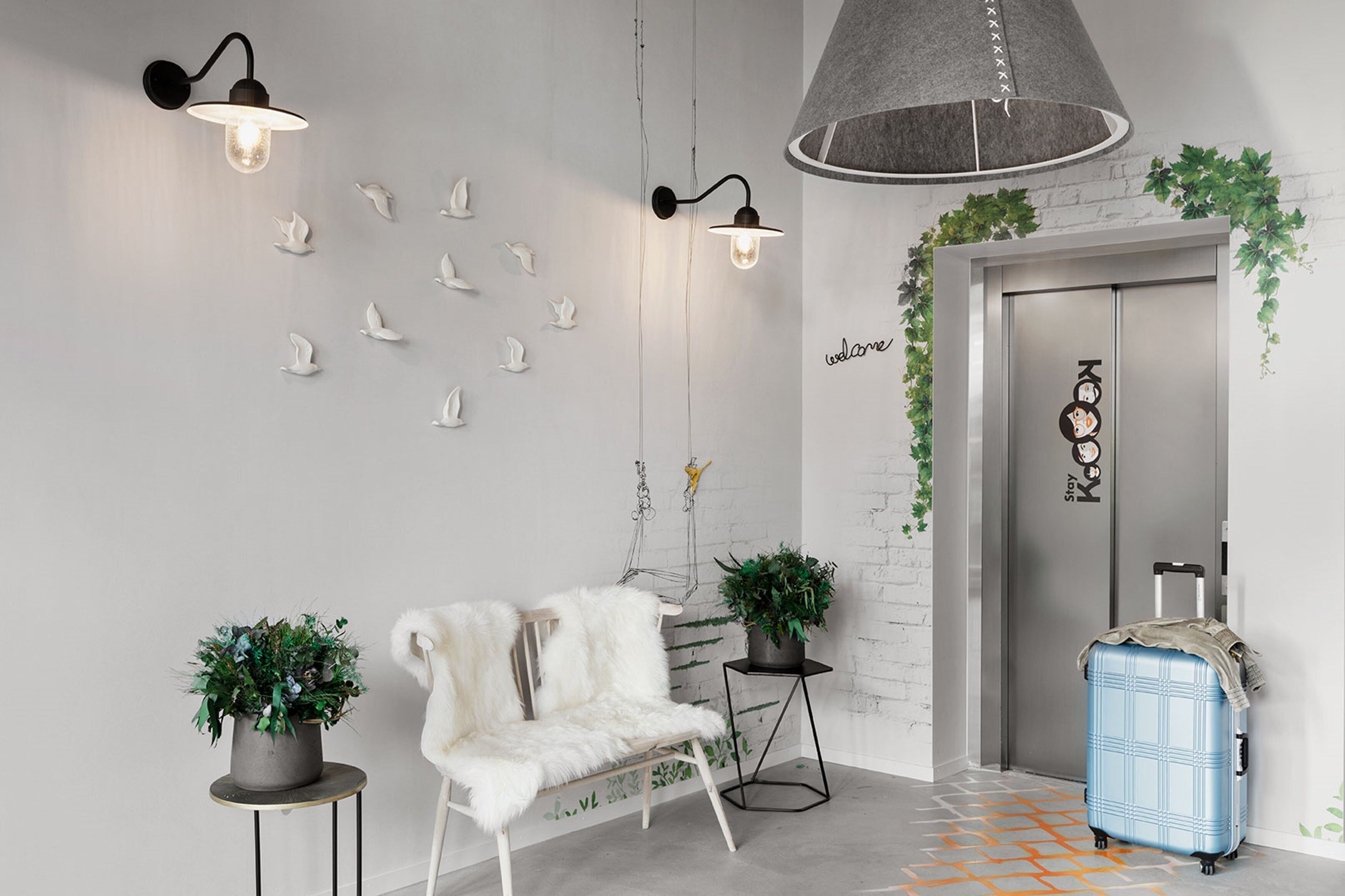 Un moderno vestíbulo con un sillón de piel blanca, dos plantas en macetas, una maleta azul e iluminación empotrada en la pared.