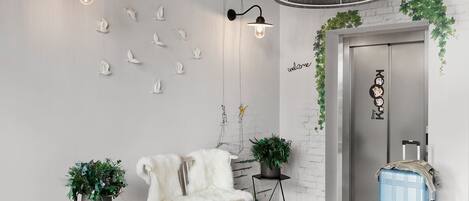 Un moderno vestíbulo con un sillón de piel blanca, dos plantas en macetas, una maleta azul e iluminación empotrada en la pared.